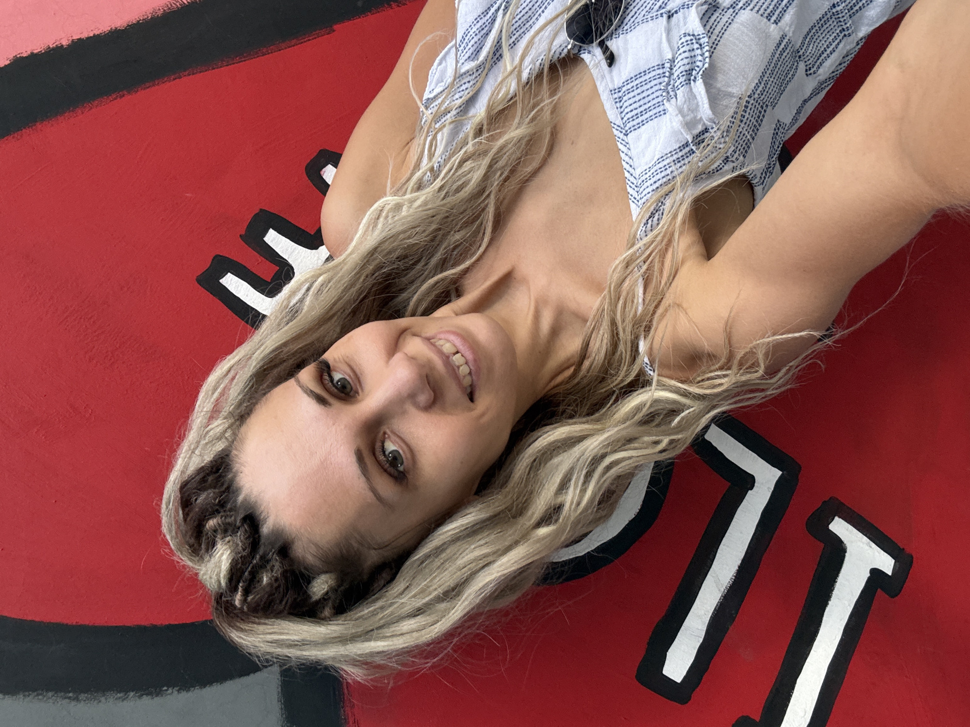 A blonde woman with braids and wavy hair is lying on a red surface with black and white graphic elements. She is looking at the camera and smiling. She is wearing a white and blue striped top. The background appears to be a mural. There is no information about the city of Rusanovo, Russia in the image.