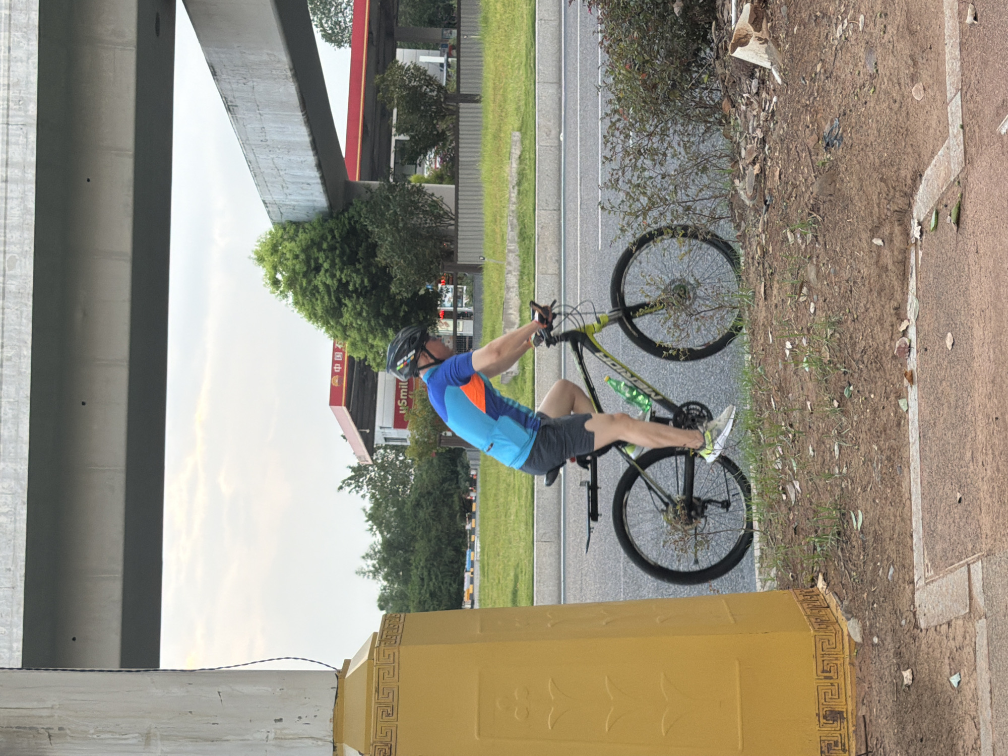 An adult male is riding a black and green mountain bike on a paved road in Jiangjin, China. The man is wearing a blue and orange cycling jersey, dark shorts, a black helmet, and white athletic shoes. He is looking forward and appears to be pedaling. The bicycle has a water bottle attached to the frame. In the background, there is a yellow pillar with decorative carvings on the left, a grassy median, and a gas station with a red canopy. An elevated highway structure is visible overhead. The sky is overcast. The scene depicts an outdoor recreational activity in an urban environment.