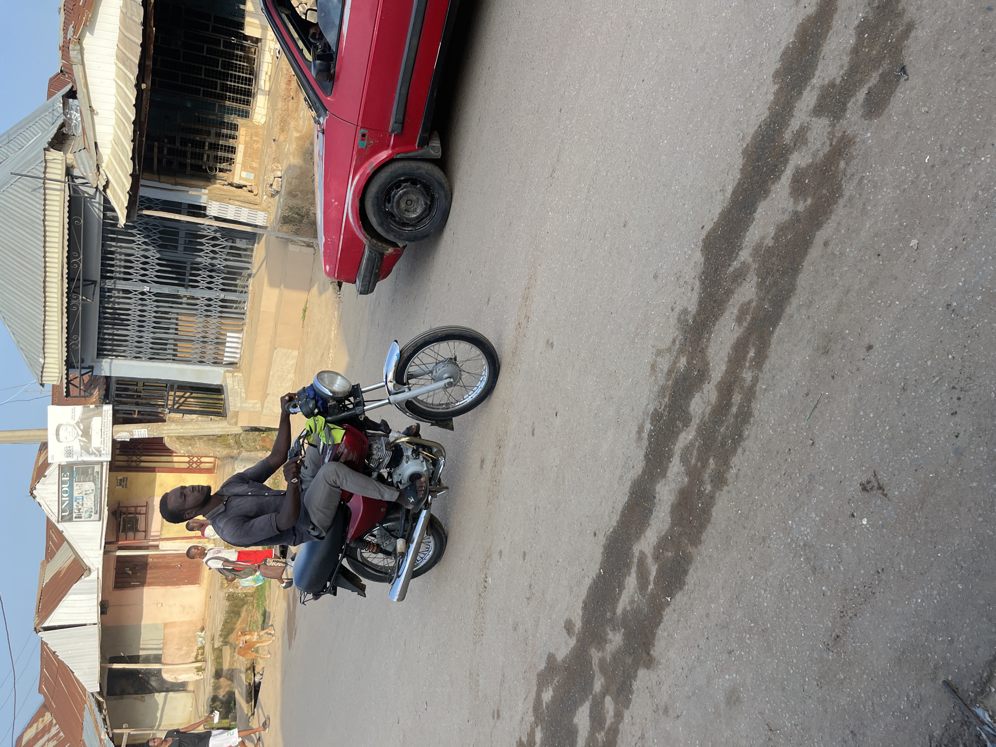 In an outdoor, urban setting resembling Abuja, Nigeria, a man is riding a red motorcycle. He is wearing a dark grey short-sleeved shirt and grey trousers, and black sandals with a white logo. The motorcycle has a black seat, a chrome exhaust pipe, and a blue headlight. To the right of the motorcycle, a red car is partially visible. In the background, there are buildings with corrugated iron roofs. A few other people and a dog are also present in the background. The ground is paved with asphalt and shows signs of wet patches. The scene appears to be a street or road in a developing urban area.