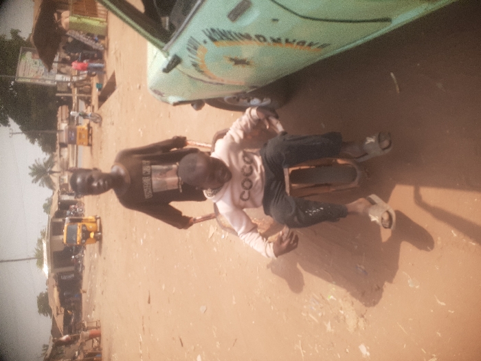 The image captures two young males on an unpaved, reddish-brown dirt surface in Gwadanye, Nigeria, under bright sunlight. A taller boy, dressed in a dark long-sleeved shirt and dark trousers, stands behind a custom-built, single-wheeled wooden cart, gripping its wooden handles. He is looking towards the camera. Seated within the wooden cart is a shorter boy, wearing a light-colored long-sleeved hoodie with "COCO" printed on the front, dark trousers, and light-colored slide sandals; he is looking downwards. To the immediate right, the front fender and door portion of a light green vehicle with faded white lettering on its side are visible. The background reveals a semi-urban landscape with various single-story structures, some featuring corrugated metal roofs. Two yellow commercial tricycles (keke Napeps) are parked further back, one on the left and one on the right. Additional indistinct figures and a palm tree are also present in the middle-ground, with utility poles or lines extending overhead.
