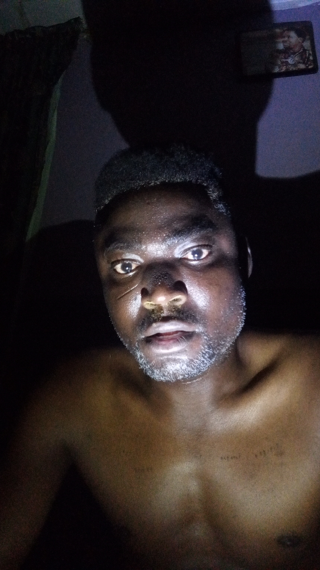 A close-up, low-light photograph shows a dark-skinned, shirtless man with a salt-and-pepper beard. He has a scar on his face, near his nose. The background is dimly lit, revealing a curtain and a framed picture on the wall. The location, Sabon Tasha, Nigeria, is not explicitly verifiable within the image alone.