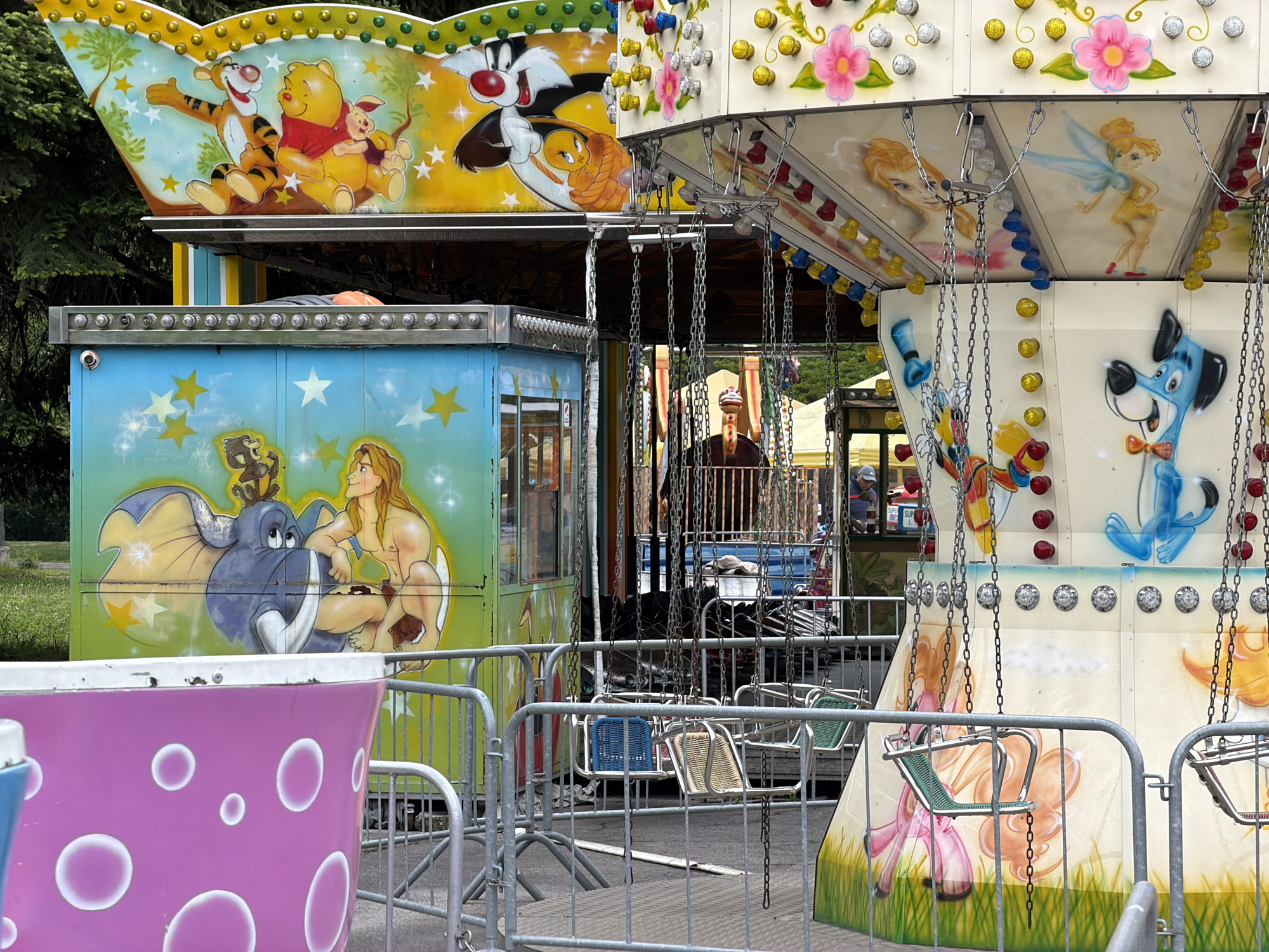 The image shows a fairground attraction, likely in Sofia, Bulgaria. The attraction features a carousel-style ride with chairs suspended by chains. The ride is decorated with cartoon characters, including Winnie the Pooh, Tigger, Sylvester the Cat, Tweety Bird, Tarzan, and other generic animated figures. The decorations appear hand-painted. The carousel is surrounded by metal barriers. A person is standing inside the carousel's operator booth. In the foreground, a section of a ride vehicle, possibly a teacup ride, is visible, painted pink with white circles. The fairground is in an outdoor setting, with trees in the background.
