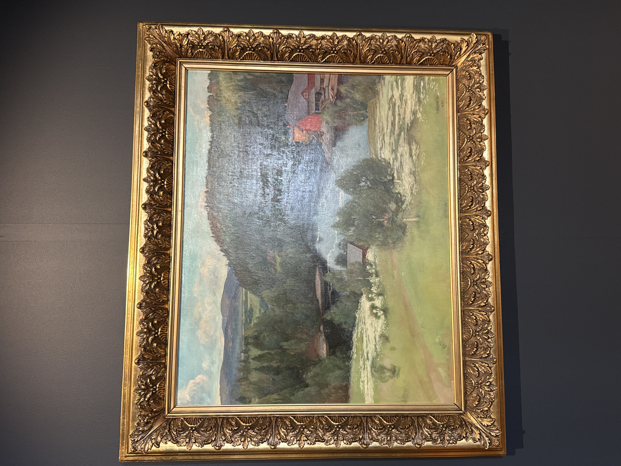 The image shows a painting in a golden, ornate frame, hanging on a dark gray wall. The painting itself depicts a landscape scene. In the foreground, there's a body of water with reflections, possibly a river or lake, with dappled sunlight on its surface. To the right, a grassy area slopes down towards the water, with a path winding through it. In the mid-ground, there are trees, and further back, on a hillside covered in dense forest, are a couple of buildings with red roofs, suggesting a rural or mountainous settlement. The background shows a continuation of the forested hillside and a glimpse of what might be mountains or distant clouds under a light blue sky.
The painting appears to be rendered in an Impressionistic style, with visible brushstrokes and a focus on capturing the play of light and color. The overall mood is serene and idyllic, suggesting a peaceful natural setting. There are no visible people in the painting, but the presence of buildings implies human habitation. The lighting suggests it could be daytime, with the sun casting shadows and highlighting various parts of the landscape.
On the right side of the painting, within the canvas, there is visible text. It appears to be a signature and a date. The signature reads "E. KAINEN" and the date appears to be "1898".