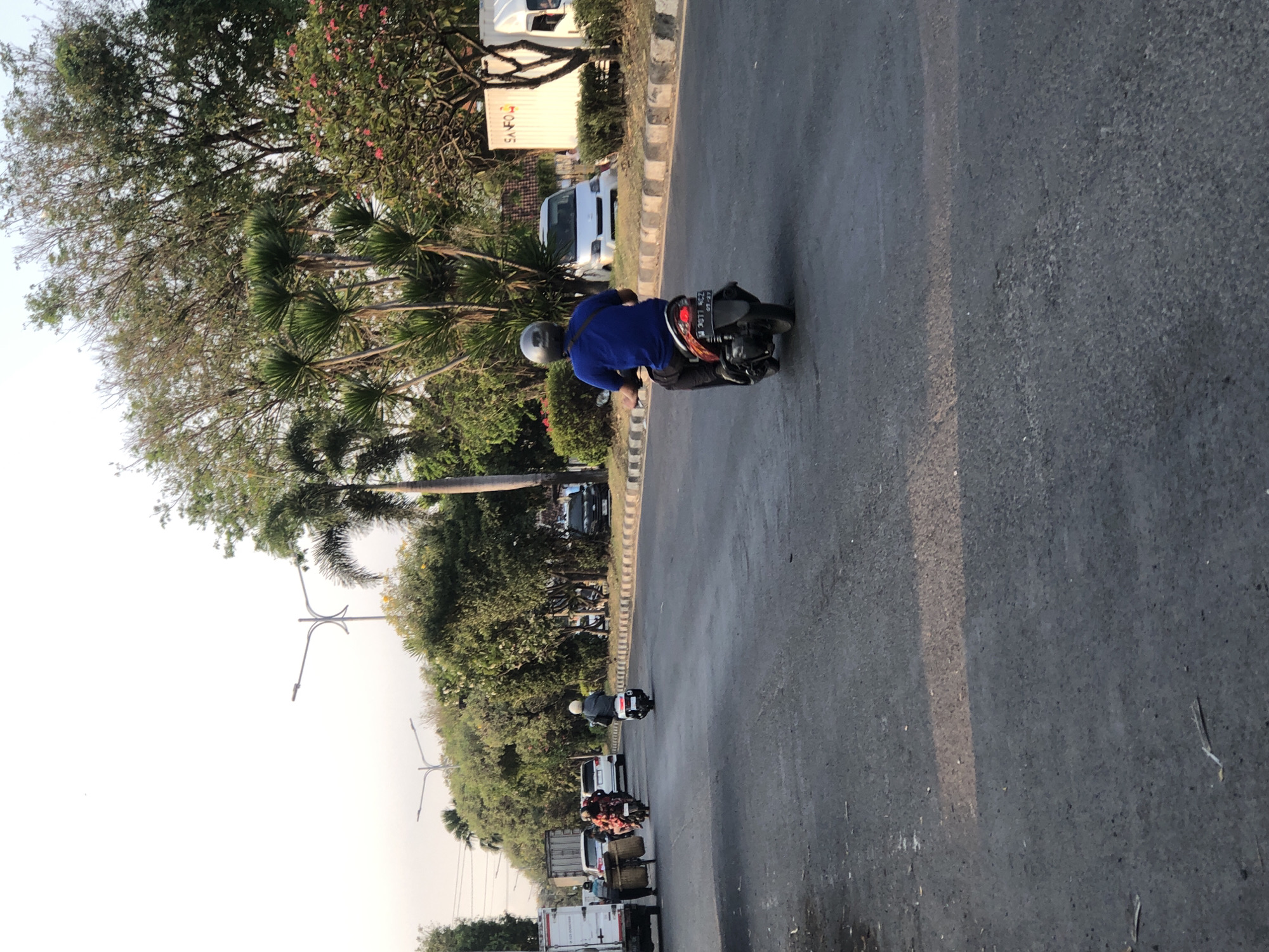 A man wearing a blue shirt and a helmet is riding a motorcycle down a paved road in the city of Ngaban, Indonesia. The road is lined with trees and bushes on both sides. Several other vehicles are visible in the background, including cars, trucks, and other motorcycles. The sky is clear and the sun is shining. The man appears to be the primary subject of the image, as he is in the foreground and in focus.