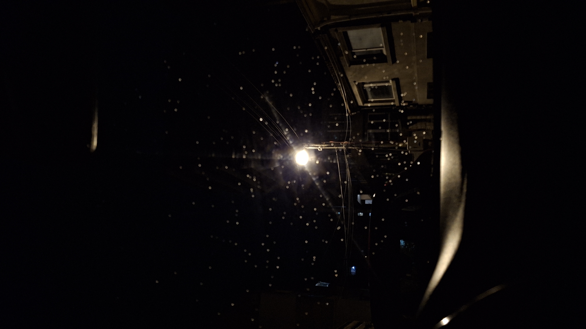 A low-angle shot at night captures a streetlamp, its bright light obscured by rain or insects, creating numerous small, blurred circles of light. Electrical wires radiate from the lamp. Above the lamp, a weathered, concrete overhang is visible. The foreground is heavily obscured by dark forms on both sides, possibly parts of a vehicle's interior. The location cannot be verified without additional context or information.