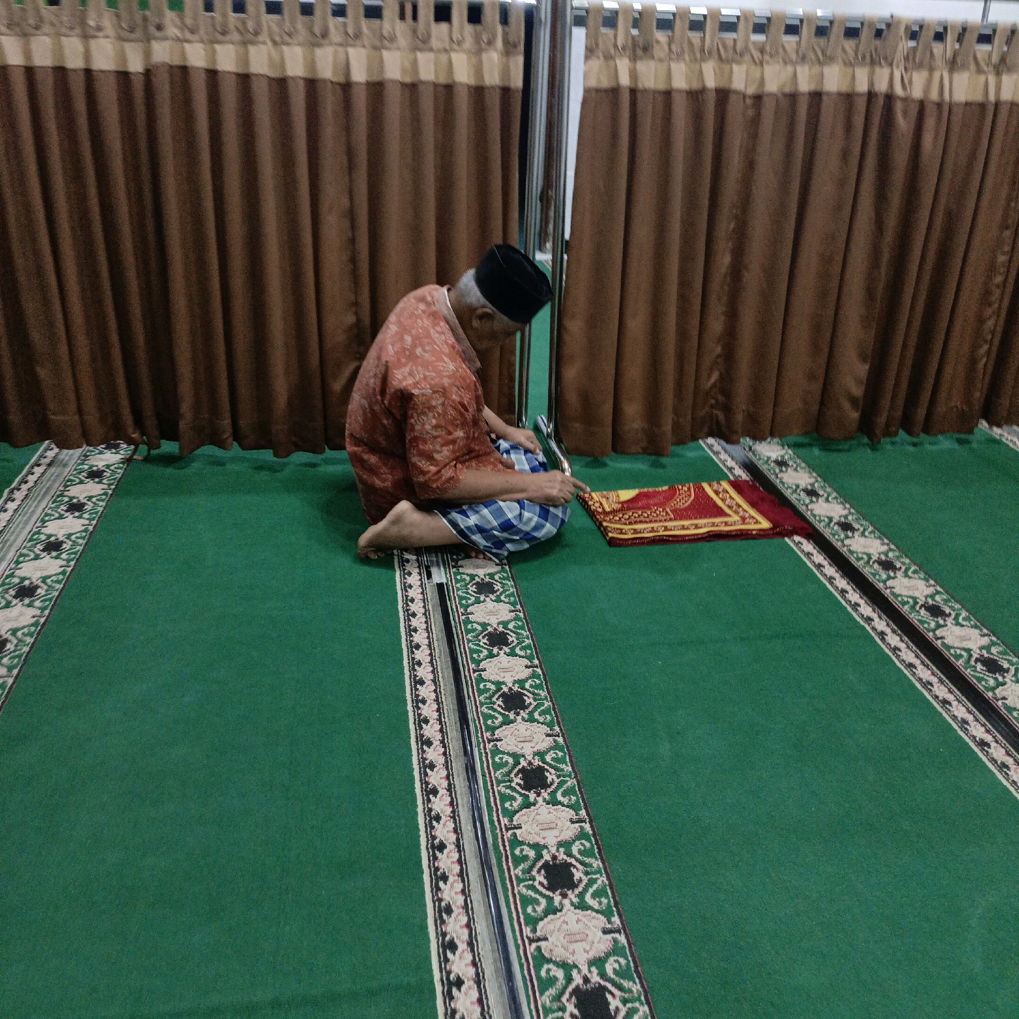 An elderly man wearing a black peci cap and a patterned orange shirt sits cross-legged on a green carpet with decorative borders. He is facing a folded red and yellow mat. The man's left hand is resting on his lap, and his right hand is reaching towards the mat. He is positioned in what appears to be a prayer room or mosque, with brown curtains and a metal railing in the background. The carpet is laid out in rows, suggesting a communal space. The scene is indoors, with artificial lighting contributing to the overall ambiance. The image is taken from a slightly elevated angle, looking down on the man and the surrounding space. The overall impression is one of quiet devotion and religious practice. This scene is likely in Salatiga, Indonesia, given the common use of such carpets and prayer mats in mosques and prayer spaces across the country.