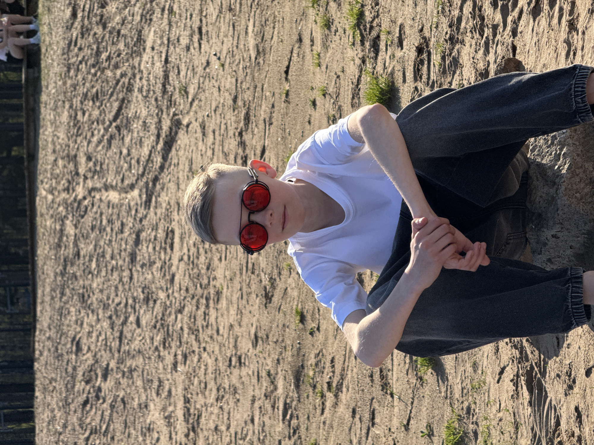 A young person is sitting cross-legged on sandy ground. They wear a white t-shirt, dark gray jeans, and round red sunglasses. The background consists of sand with scattered grass. A blurry dark object is on the left edge of the frame. The location is Chaikovsky, Russia.