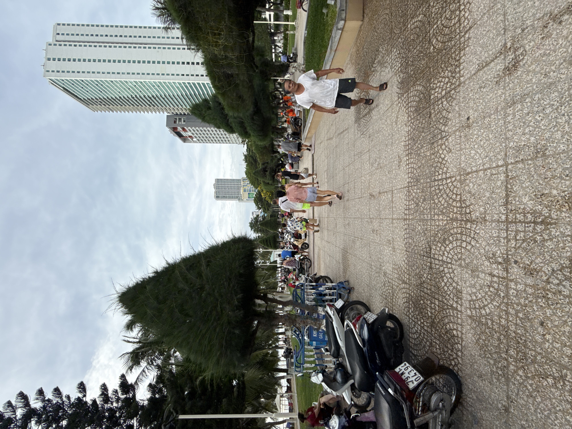 The image depicts a scene on a paved walkway in Nha Trang, Vietnam, likely near a beach due to the presence of palm trees and a cloudy sky suggesting a warm climate. In the foreground, a man in a white t-shirt and dark shorts walks towards the viewer, his expression neutral. Behind him, other pedestrians are walking along the path, some in casual summer attire. A row of parked motorcycles lines the left side of the walkway, suggesting a common mode of transportation in the area. The pathway itself is paved with decorative concrete tiles. In the background, modern high-rise buildings stand tall against the sky, indicating an urban setting. Lush green trees and grass areas are visible, providing a contrast to the concrete structures. The overall atmosphere appears to be one of everyday activity in a coastal city.