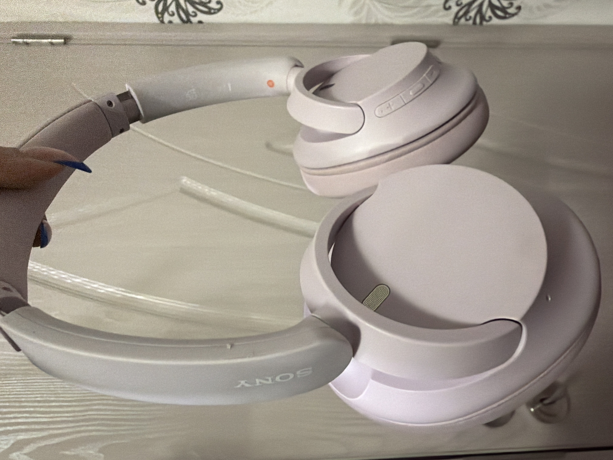 A pair of light pink or lavender Sony brand over-ear headphones are depicted, held by a human hand. The headphones feature a sleek, integrated design with the 'SONY' logo embossed vertically on the outer left headband. The right earcup displays multiple physical control buttons and a small orange indicator light near its hinge. A silver-colored mesh-like component is visible on the interior side of the left earcup. The hand holding the headphones at the top center has medium brown skin and fingernails painted in a dark blue hue. The background comprises a light-colored door or wall with vertical paneling on the left, and a section of light-colored wallpaper featuring dark, ornate patterns on the right. A metallic door hinge is partially visible on the right side, and a metallic door handle with a lock cylinder is present on the lower left. The scene is located indoors in Pazardzhik, Bulgaria.