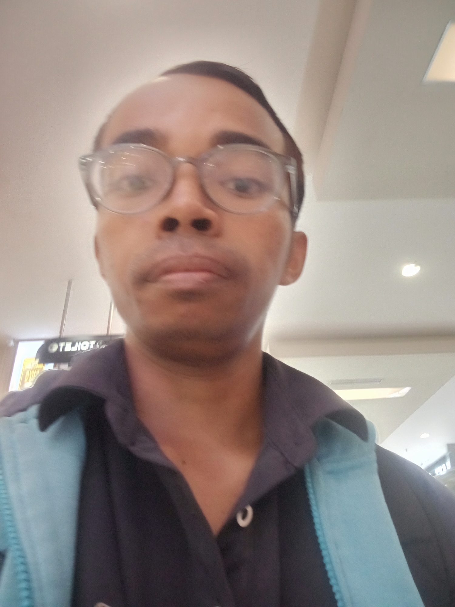A man with glasses is taking a selfie. He is wearing a dark collared shirt and a light blue jacket. In the background, there is a sign that reads "TOILETS" in reverse. The environment appears to be an indoor public space with overhead lighting. The image quality is low, and the man's face is slightly blurred.