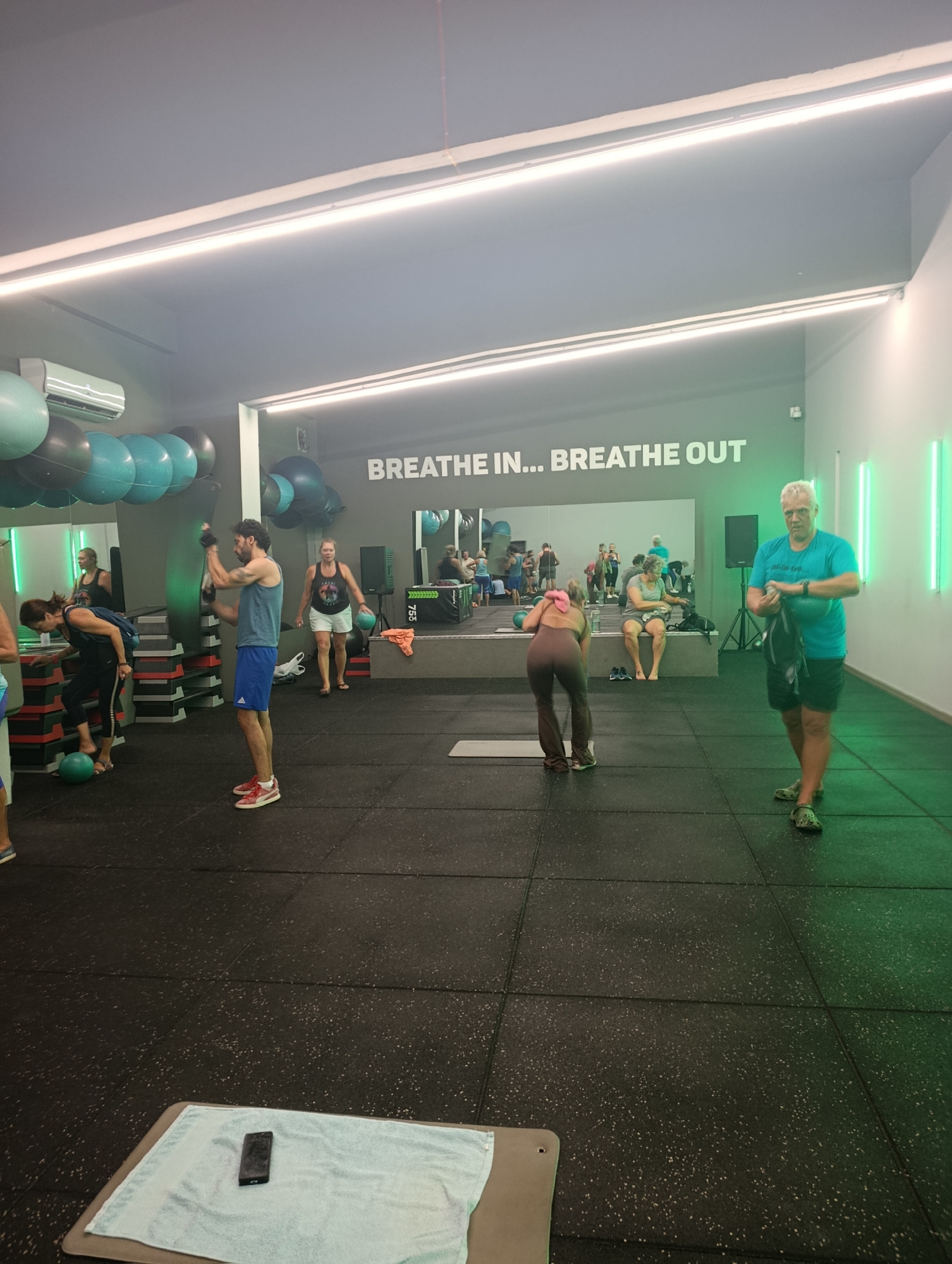 The image depicts the interior of a gym. In the background, a motivational phrase, "BREATHE IN... BREATHE OUT," is illuminated on a wall. Several people are engaged in various fitness activities. In the foreground, a man in a blue tank top and shorts stands with his arms crossed, holding a black bag. To his right, another man in a turquoise t-shirt and shorts also stands holding a black bag. The gym floor is black rubber matting. The environment is lit by overhead lights and green neon lights on the right wall. In the foreground, a towel with a phone on it rests on a mat. The gym appears to be located in Setúbal, Portugal, given the context of the request.