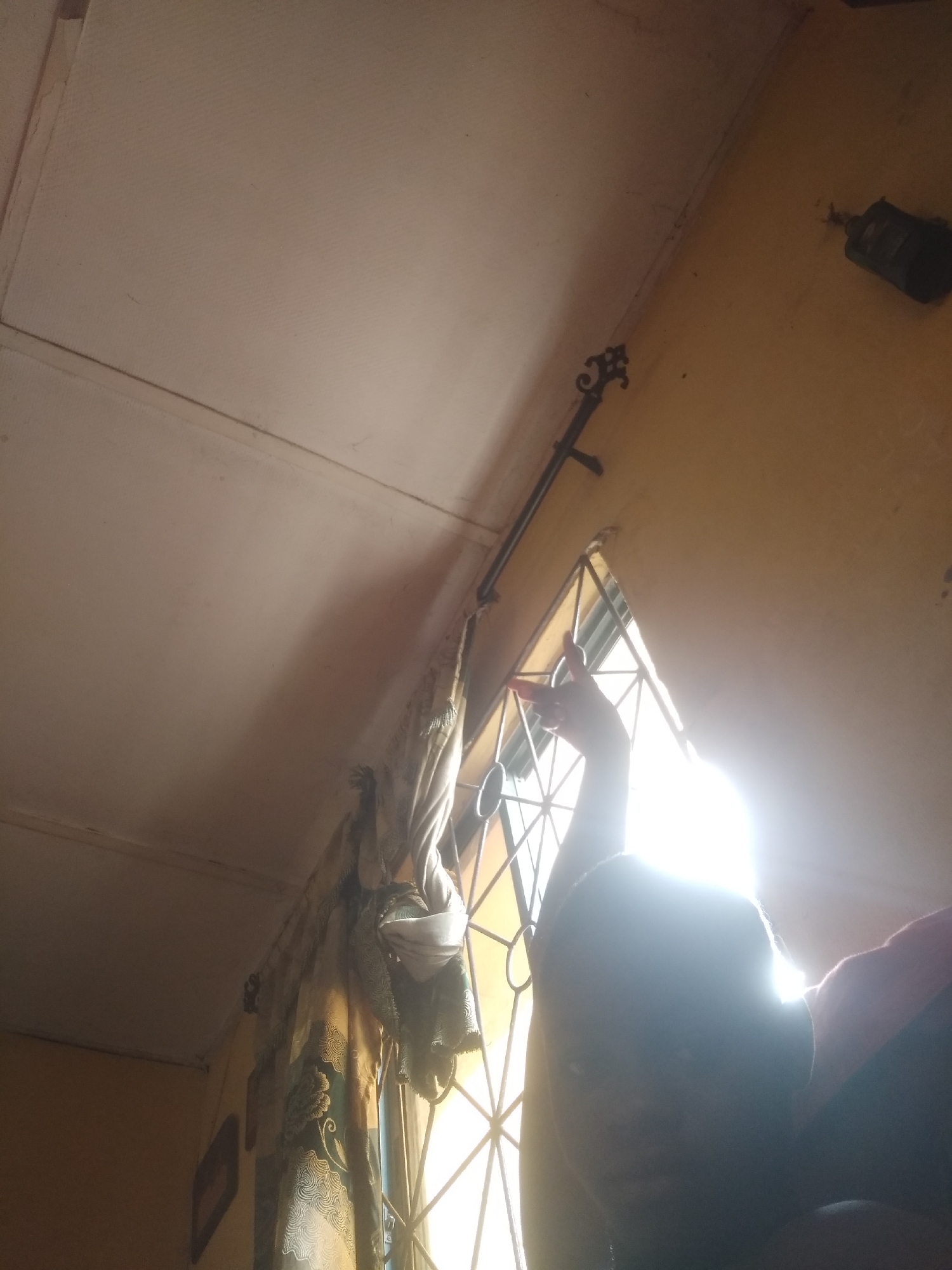 The image appears to be taken indoors, possibly in a residence in Shangisha, Nigeria, given the contextual request. The viewpoint is looking upwards towards a window. The window is a geometrical pattern of crossed bars. Sunlight is intensely shining through the window, which makes it difficult to see details directly in front of the window. The subject in the foreground is silhouetted against the bright light. A hand is lifted up toward the window. To the left of the window, there is a curtain rod with a decorative end piece. Curtains are draped on the rod, some of which hang down. To the upper right of the window, there is a wall, and near the top right corner of the wall is an object that is not easily discernible due to the image quality. The ceiling is visible, with a distinct seam line running along its surface.