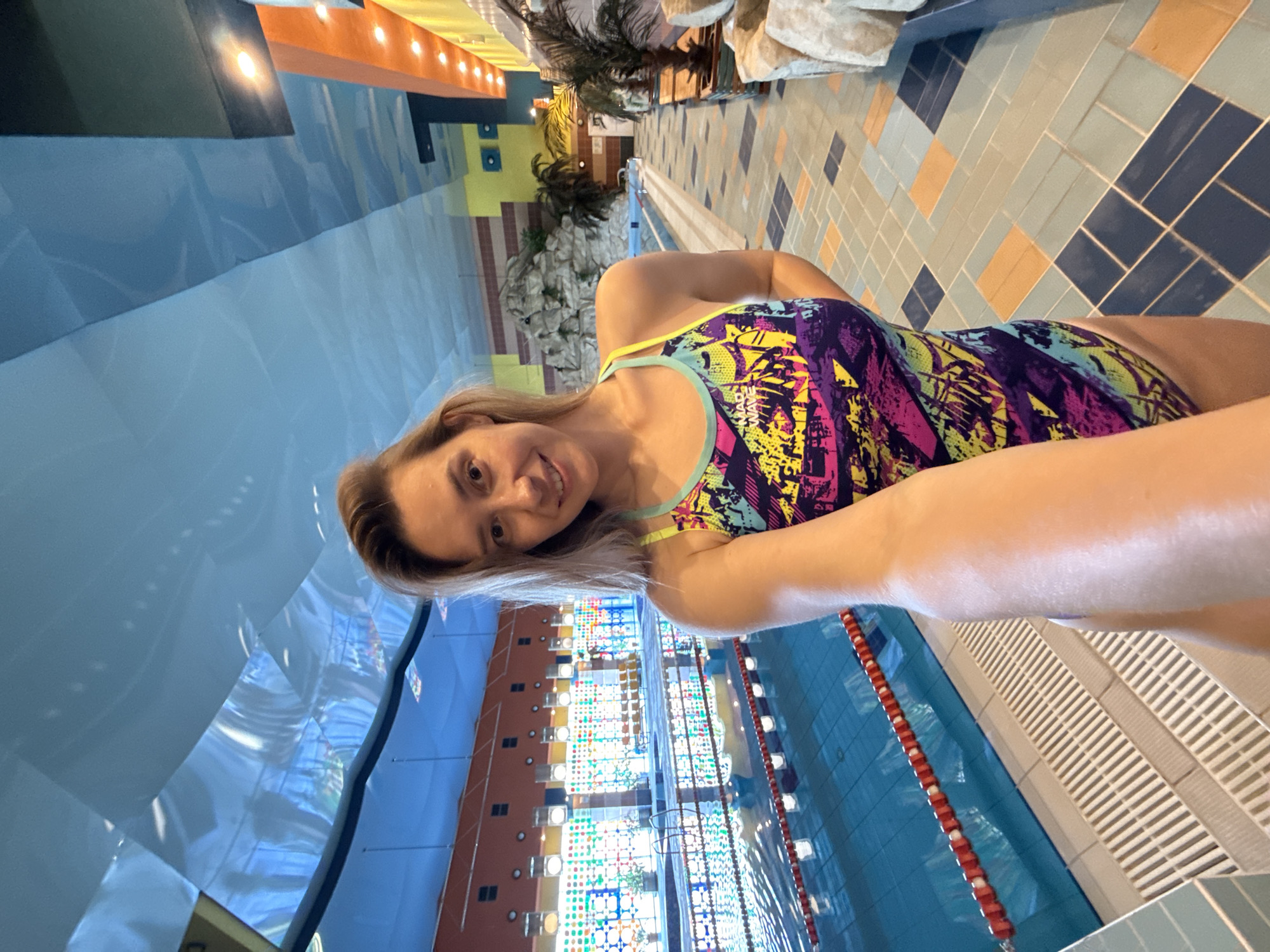 A woman is taking a selfie in a swimming pool area. She is wearing a colorful one-piece swimsuit. The pool is visible in the background, with lane lines and a blue bottom. The surrounding area has tiled floors, artificial rock formations, and palm trees. The ceiling is blue with interesting lighting. This appears to be an indoor swimming complex, possibly a public facility. The image does not contain any information that allows for identification of its specific location in Chaikovsky, Russia.