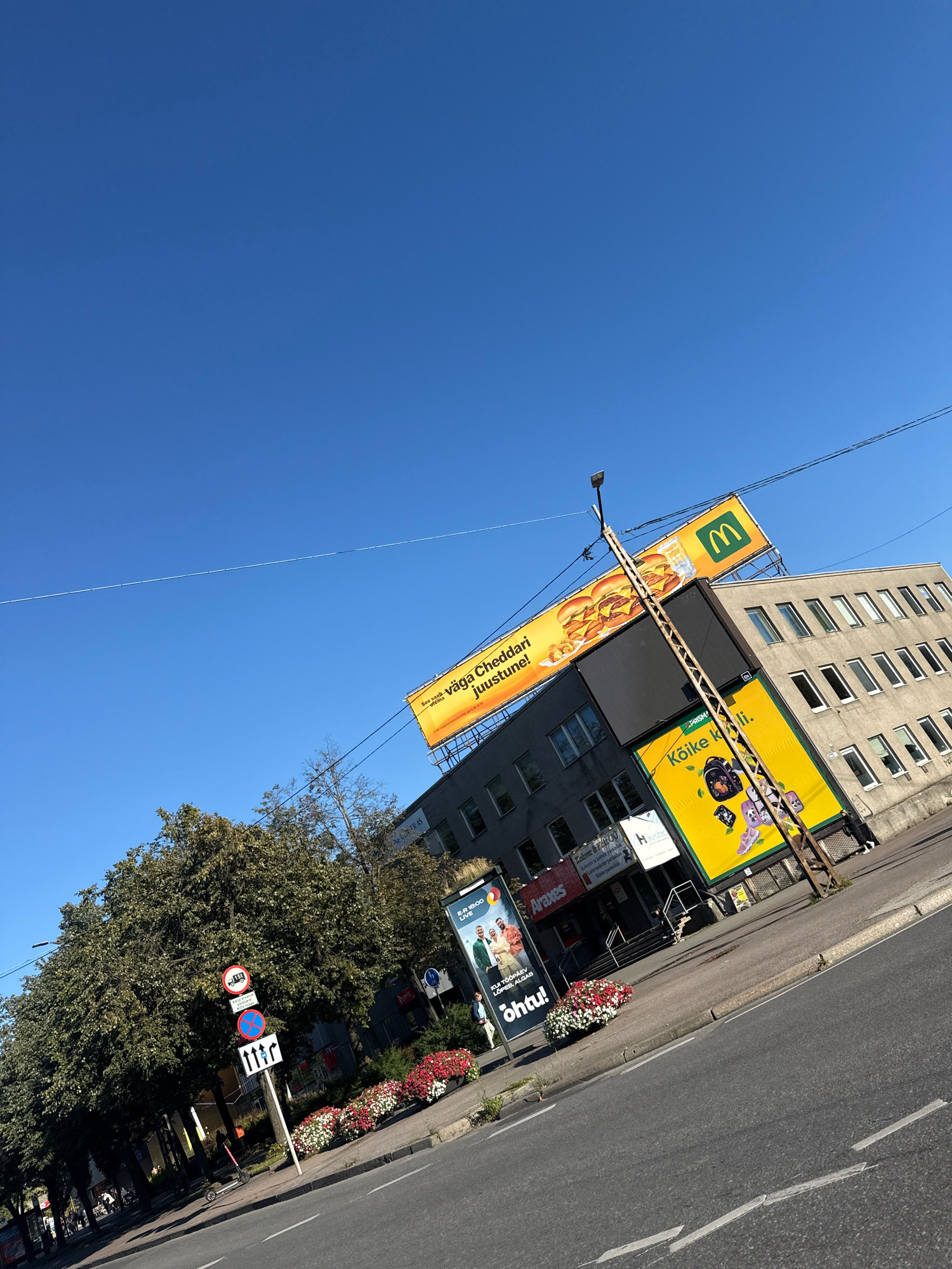 In Tallinn, Estonia, a bright blue sky serves as the backdrop for a sunlit street scene. On the right, a modern, multi-story building with numerous windows stands tall. Prominently displayed on the building's facade are large billboards advertising McDonald's and a product featuring children's school supplies. A sturdy metal pole supporting power lines runs vertically in front of these billboards. To the left, a dense, green tree provides shade over a sidewalk lined with colorful flower beds. Various traffic signs, including a prohibition sign and directional arrows, are visible near the trees. A red electric scooter is parked on the sidewalk. The street in the foreground shows painted lane markings. People are present on the sidewalks, and a few vehicles are visible in the distance. The overall atmosphere is that of a typical urban environment on a clear day.