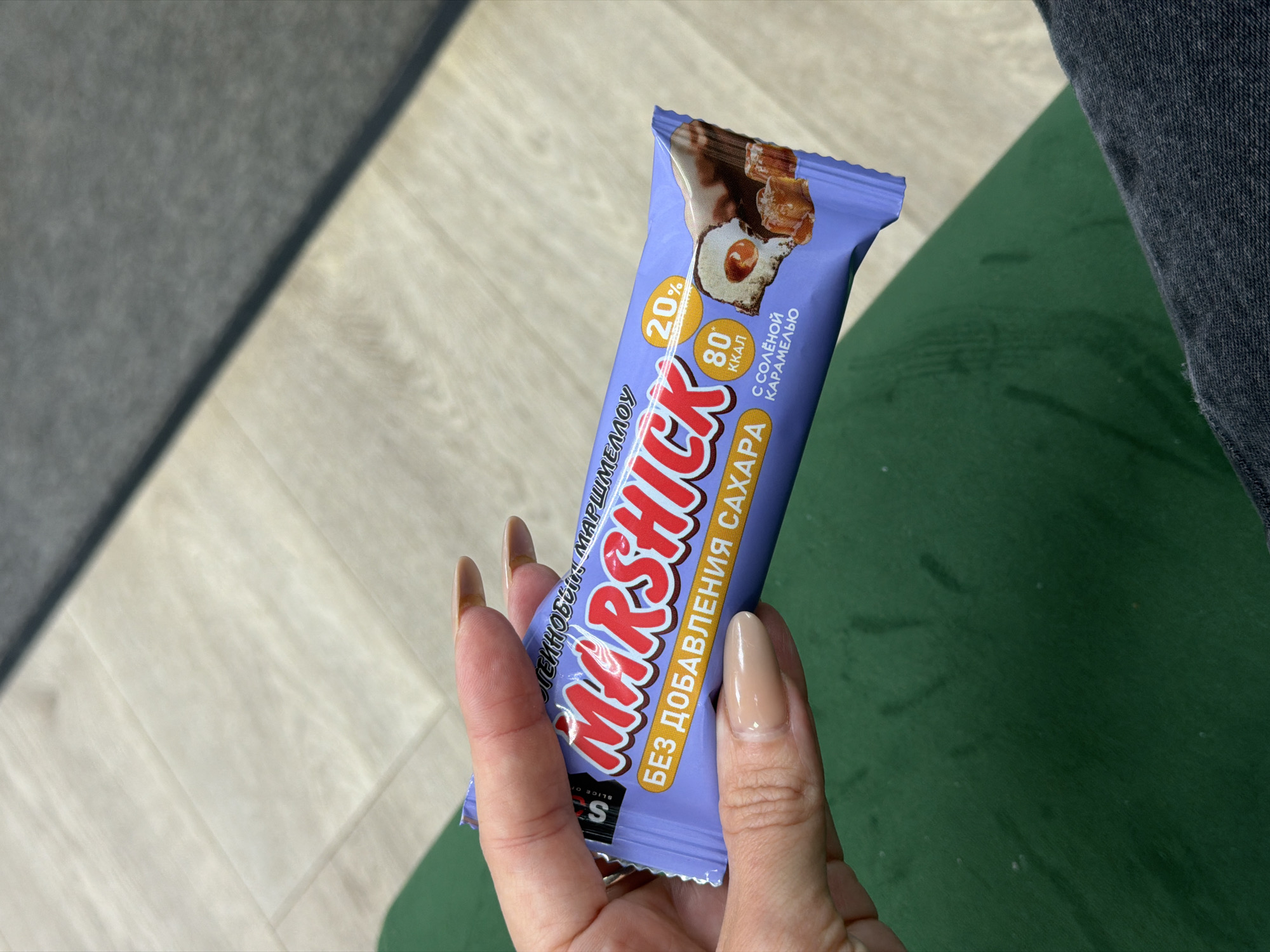 A person's hand with neatly manicured nails holds a purple candy bar wrapper. The wrapper features the brand name "Marshick" in large red letters, along with text in Russian indicating it is a marshmallow candy bar with no added sugar, 80 kcal, and salted caramel. A cross-section of the candy bar is depicted, showing marshmallow and caramel filling. The background consists of a light-colored floor, possibly laminate or linoleum, and a dark gray fabric, possibly clothing. The lighting suggests an indoor setting, possibly a public space or an office in the city of Perm, Russia.