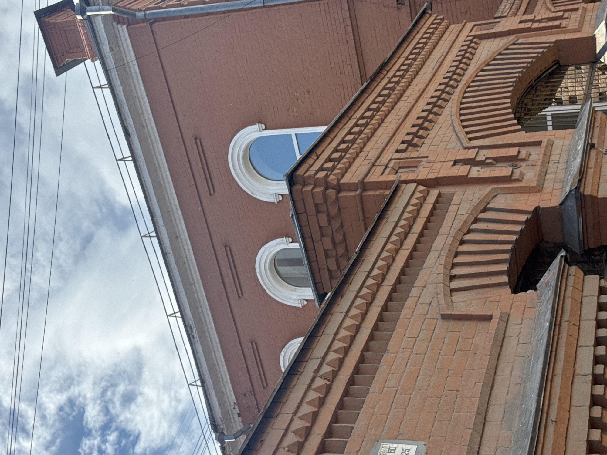 The image shows the facade of a brick building in Yekaterinburg, Russia. The building has red brick walls and features several arched windows with white decorative trim. The windows are of varying sizes, with some being semicircular and others more elongated. The architecture appears to be of an older style, possibly industrial or institutional. There are visible details of the brickwork, including horizontal and vertical decorative elements. The sky is visible in the upper portion of the image, with scattered clouds. Electrical wires are strung across the sky above the building. The photograph is taken from a low angle, looking up at the building.