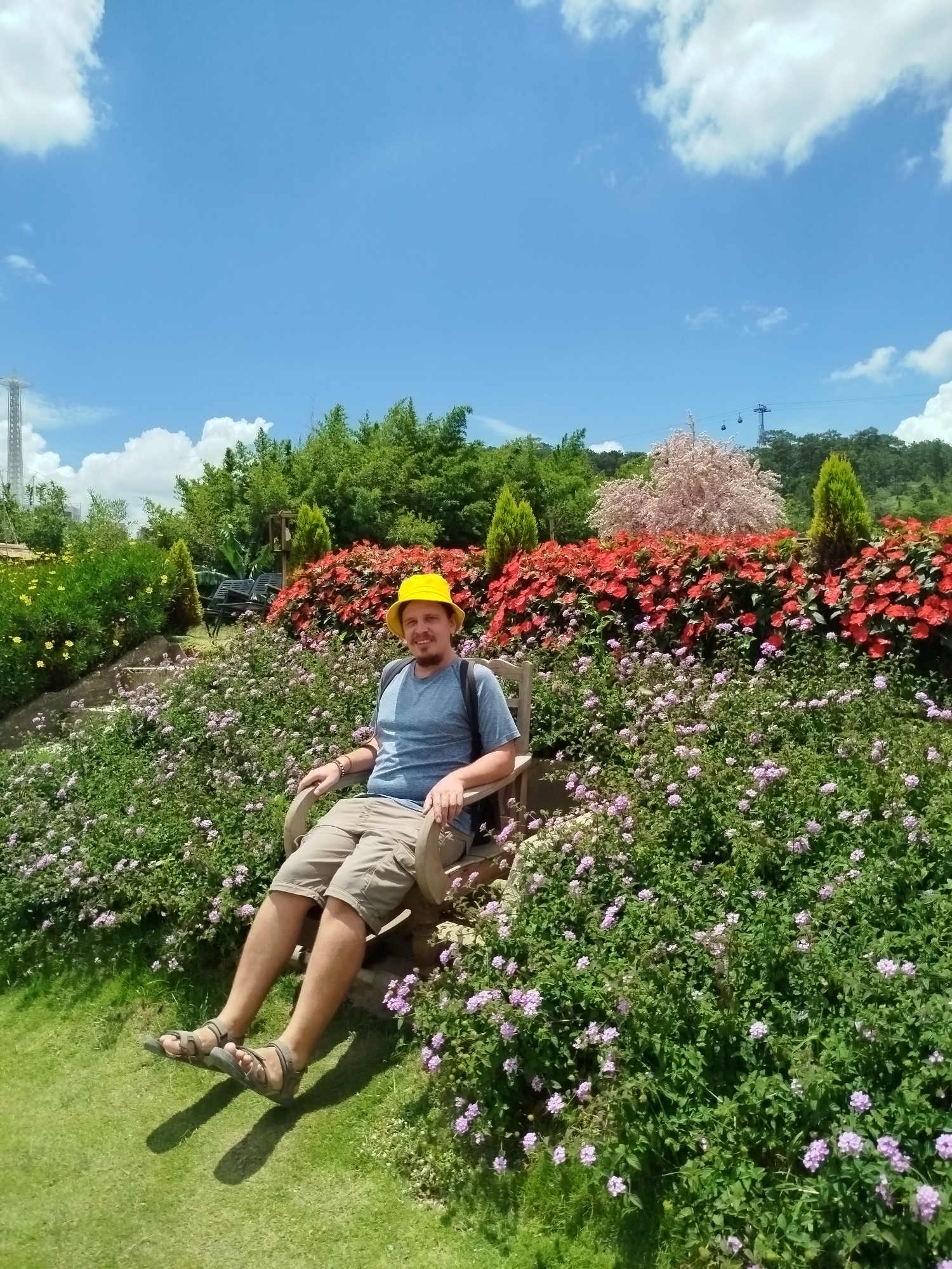 A man wearing a yellow hat and a blue t-shirt is sitting on a wooden chair in a garden. He is wearing khaki shorts and sandals. The garden is full of flowers and greenery. In the background, there are trees and a blue sky with clouds. The scene appears to be in a park or botanical garden. It is possible this is the Datanla High Rope Course, a popular tourist attraction in Da Lat, Vietnam, known for its beautiful gardens and outdoor activities.