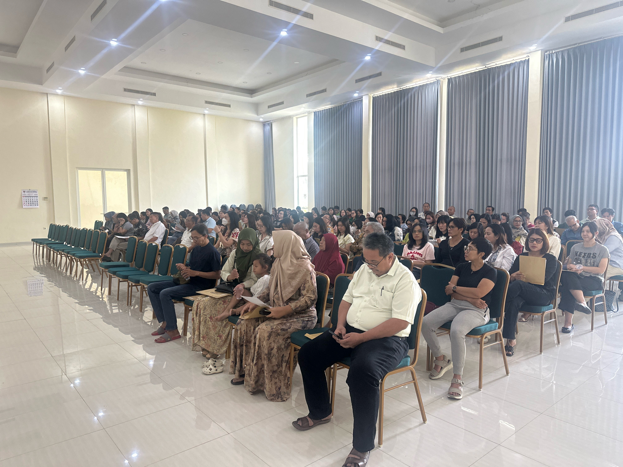 The image depicts an indoor event, likely a conference or a seminar, held in the city of Mojokerto, Indonesia. A large group of people, diverse in age and gender, are seated in rows of dark green chairs facing forward, suggesting they are an audience. The room is well-lit by overhead fluorescent lights and appears to have a high ceiling with a white, possibly tiled, surface. The walls are painted a pale yellow and are adorned with ventilation grilles and what appear to be large, grey curtains. The floor is tiled with large, white square tiles. In the foreground, a man in a light yellow shirt and black pants is seated, looking at his phone. To his left, a woman in a patterned dress and headscarf is looking down, while a child in a white shirt sits between her and another woman in a patterned dress. Rows of empty chairs are visible on the left side of the image, and a calendar is affixed to the wall in the far left. The overall atmosphere suggests a formal gathering.