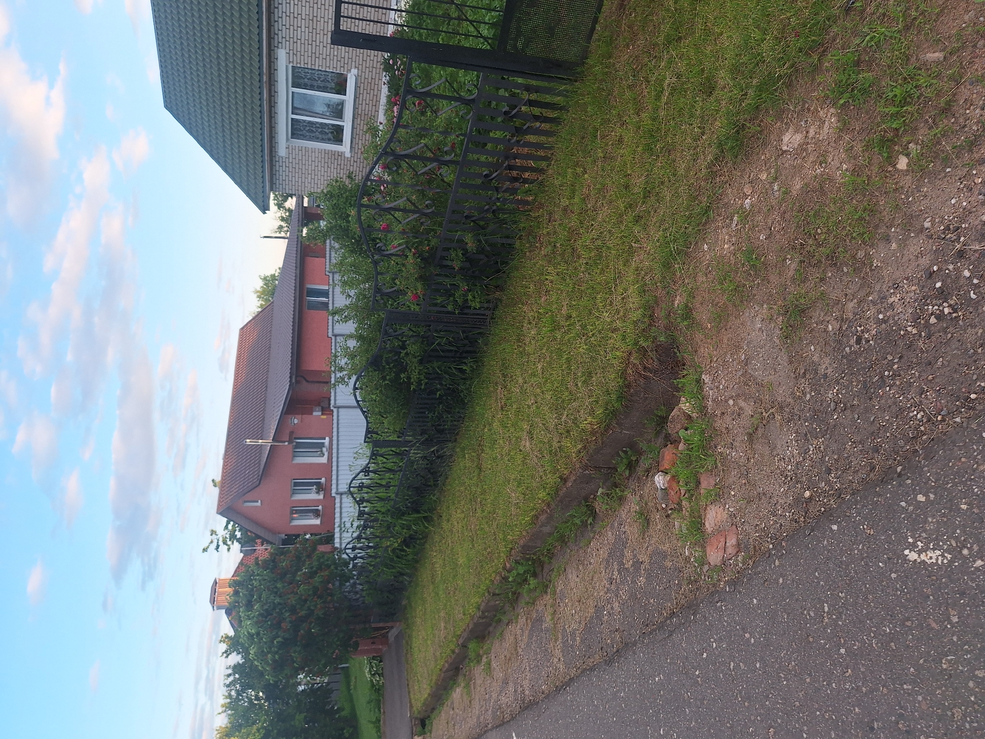 The image depicts a street-side view of two residential houses with a black metal fence in front of them, located in the village of Kamyanets, within the Kapyl District of Belarus. The house on the left is a two-story structure with a terracotta-colored roof and a salmon-pink facade. It features multiple windows and a prominent chimney. Adjacent to it is a mature tree with red berries. The house on the right is a single-story dwelling with a green, textured roof and a light-colored brick exterior. It has a large white-framed window. The fence, made of wrought iron with decorative elements, runs along the front of both properties, separating them from the road and a grassy verge. The verge is covered in well-maintained grass and bordered by a concrete curb. A paved road occupies the foreground, with loose gravel and some discarded red bricks visible on the edge of the verge. The sky above is a clear blue with scattered white clouds. The overall scene suggests a quiet, residential area.