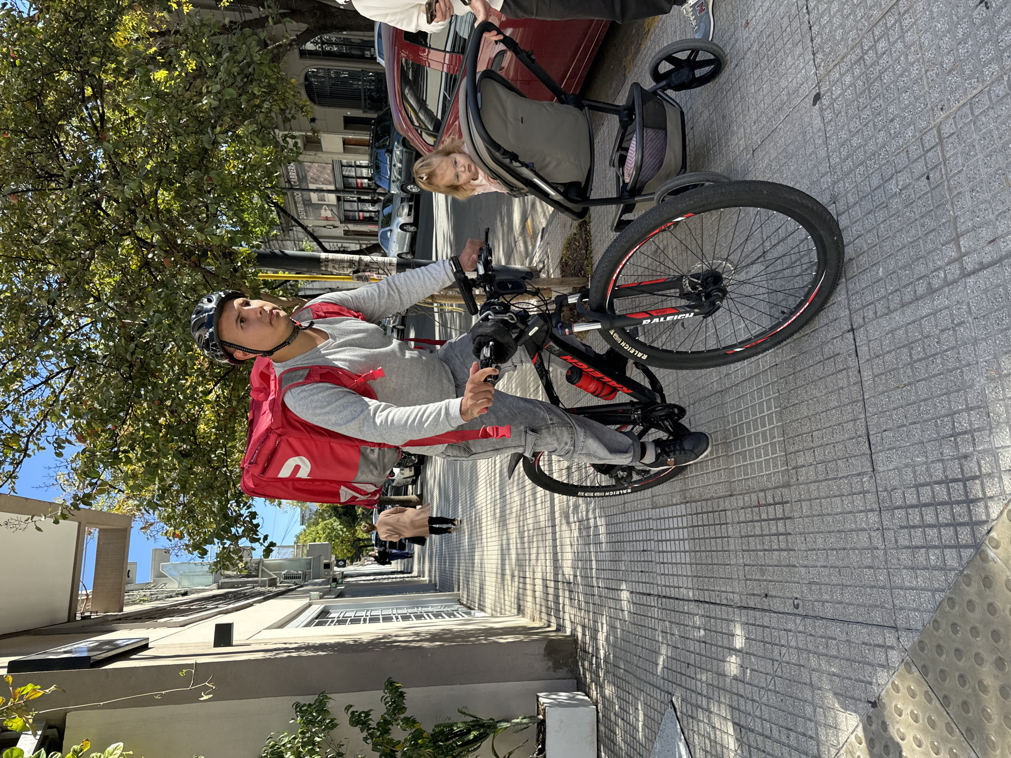 This image depicts a sunny urban street scene in Buenos Aires. In the foreground, a man, likely a delivery rider, is paused on a black and red "RALEIGH Sprint" bicycle. He wears a grey long-sleeved shirt, blue jeans, a black helmet, and a bright red delivery backpack featuring a white "U" logo, characteristic of services like Uber Eats. He holds the handlebars and looks slightly to his left.
Adjacent to the man, a woman (mostly out of frame) pushes a grey baby stroller. A young child with blonde hair is seated in the stroller, looking directly towards the camera with a neutral, curious expression.
The setting is a city sidewalk paved with grey square tiles, illuminated by strong daylight that casts clear shadows, indicating midday or afternoon. Green trees with abundant foliage line the street, providing natural shade. In the background, parked cars are visible, including a red sedan next to the stroller. Buildings with traditional architecture, including arched windows, frame the street. A prominent sign in the distance reads "DISTRIBUIDORA CABRERA" with "MEAT" below it. The overall atmosphere suggests a casual moment on a bustling city street.