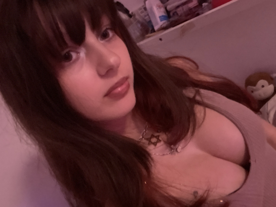 A woman with brown hair and bangs poses in a selfie. She wears a taupe top, a silver chain, and a Star of David necklace. Her lips are glossy. In the background, items are on a shelf, and a stuffed monkey is visible behind her. The lighting has a pinkish tint.