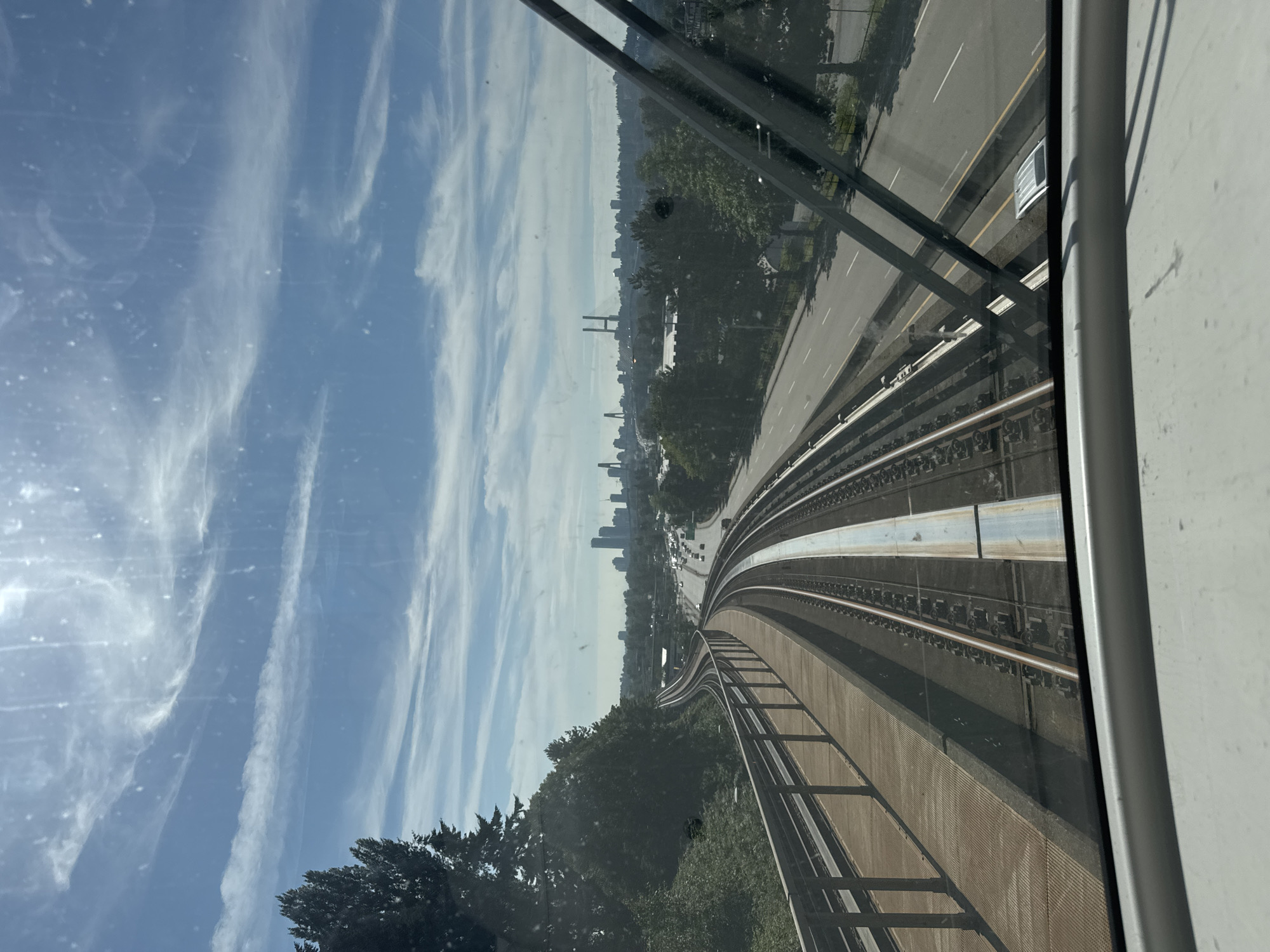 The image shows a view from a SkyTrain in Surrey, Canada, looking out towards the Vancouver skyline on a bright, sunny day with scattered clouds. The SkyTrain tracks curve to the left, with a walkway and metal railing alongside. To the right of the tracks is a multi-lane road with several cars. In the distance, the Vancouver skyline is visible, featuring tall buildings and bridges. Lush green trees and vegetation line the sides of the tracks and the road. The windshield of the SkyTrain is visible in the foreground, with wiper blades and some water spots. The scene captures a typical urban commute with a scenic backdrop.