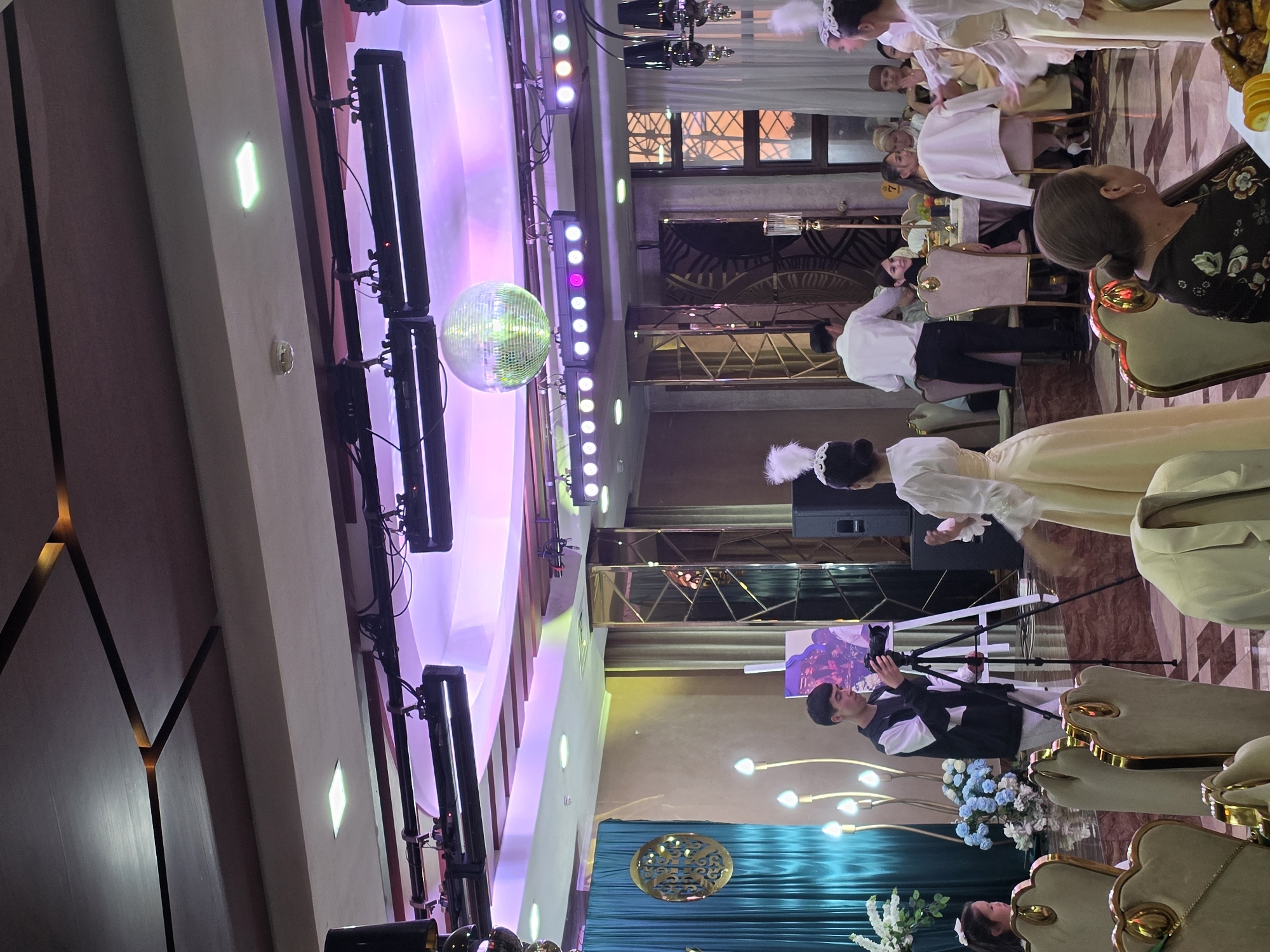 The image shows an event taking place in what appears to be a banquet hall in Bishkek City, Kyrgyzstan. A disco ball hangs from the ceiling amid stage lighting. A photographer with a camera on a tripod stands in front of a turquoise backdrop with a decorative gold emblem, possibly filming or photographing people in costume. Many people are sitting at tables, seemingly observing or participating in the event. The seating arrangements suggest a formal dinner or celebration. The ambiance is lively with colorful lighting and festive decor.