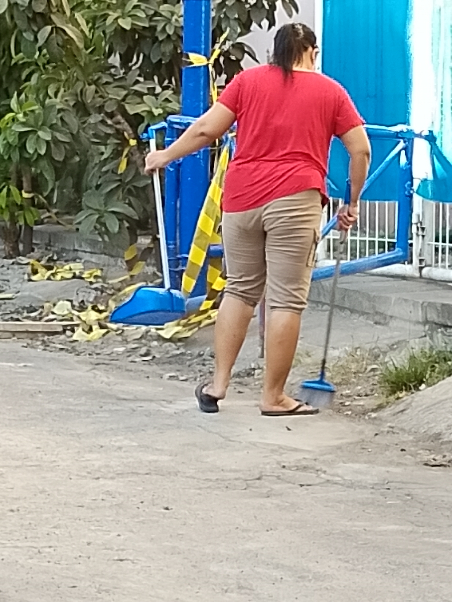 A person wearing a red t-shirt and beige capri pants is sweeping a dirt and gravel path with a blue-bristled broom. The person is also wearing black flip-flops. Behind the person, there is a blue structure with yellow and black striped caution tape. Trees with green leaves are visible to the left of the scene. The ground appears to be a mix of dirt, gravel, and possibly construction debris. The scene is outdoors and appears to be in an urban or suburban setting. The lighting suggests it is daytime. The overall impression is one of cleaning or maintenance work. The location is identified as Surabaya, Indonesia, likely an outdoor public space or a residential area undergoing minor construction or cleanup.