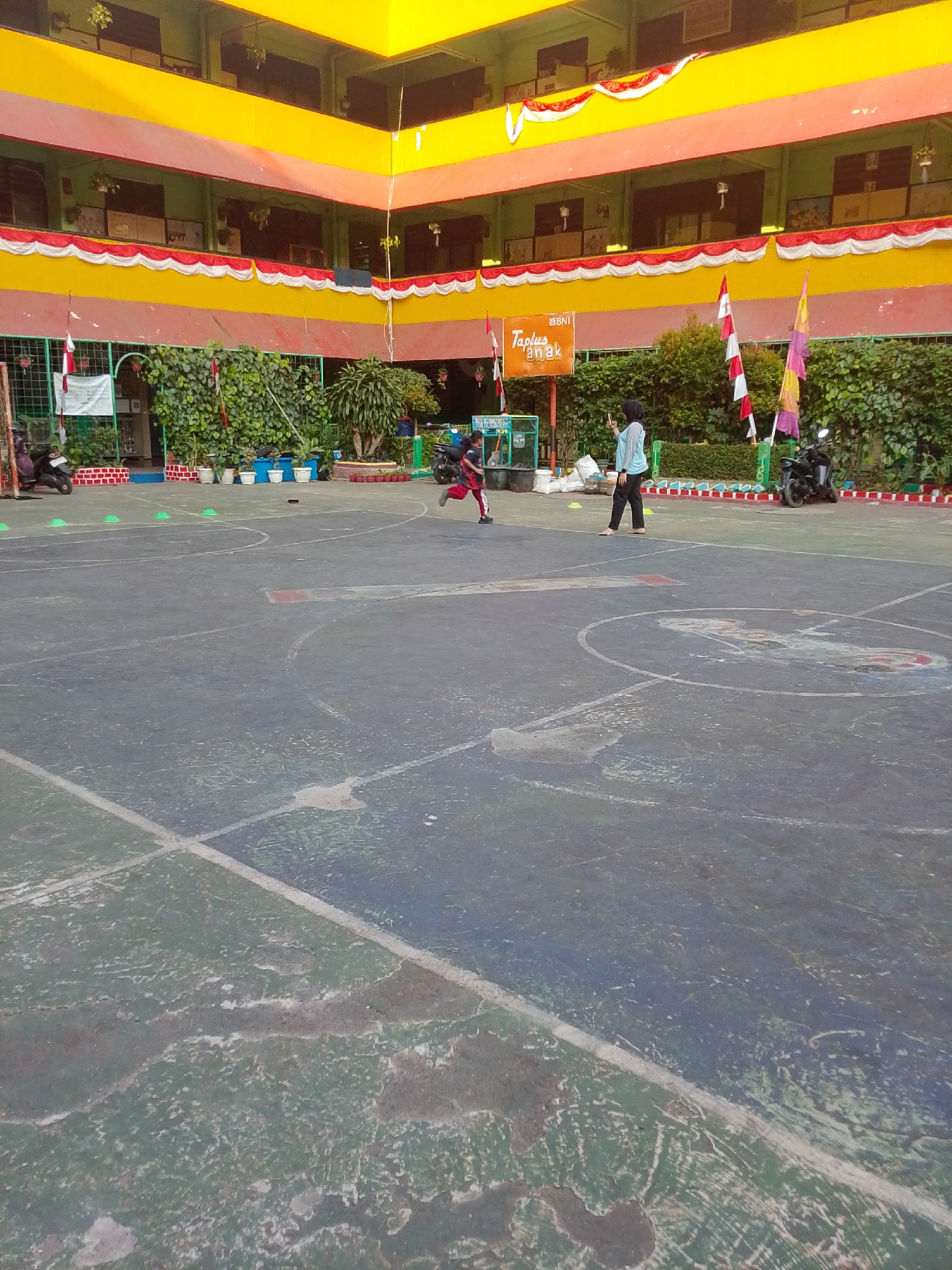 The image depicts an outdoor concrete basketball court, possibly part of a school or community center, in Central Jakarta, Indonesia. The court surface is worn and shows areas of green and grey discoloration. Faint white lines marking court boundaries are visible. In the background, a multi-story building with vibrant yellow and red trim frames the scene. A row of green plants and some potted plants are situated along the building's base. Several Indonesian flags, red and white, are displayed on poles. A dark-colored motorcycle is parked on the right side of the court.
Two individuals are present on the court. A child, wearing a red shirt and black shorts, is running in the middle of the court. Another person, wearing a light blue long-sleeved shirt and dark pants, stands to the right of the running child, holding a smartphone, possibly taking a picture. Green cones are scattered across the court, suggesting an organized activity or training session. The lighting suggests it is daytime.