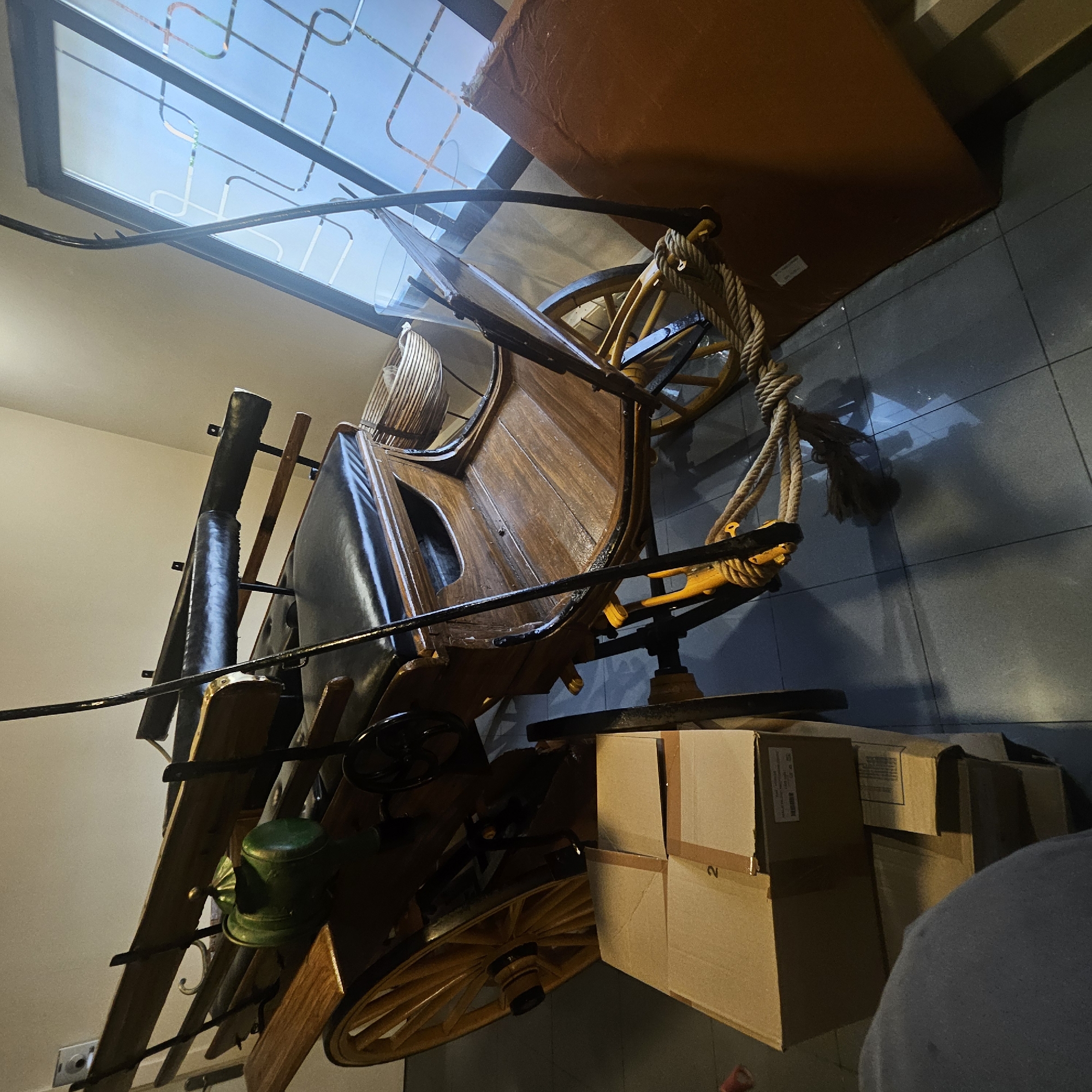 In Almadrones, Spain, a horse-drawn carriage is partially visible, resting on a tiled floor. The carriage features a wooden body with a black leather seat and a wooden passenger bench. A green, dome-shaped object is placed on the carriage. To the left of the carriage, several cardboard boxes are stacked, with a prominent "2" visible on the top box. A large, brown, flat object is propped against the right side of the carriage. The background includes a wall and a large window with a patterned design. The scene appears to be indoors, possibly in a storage area or a garage. There are no people visible in the image.