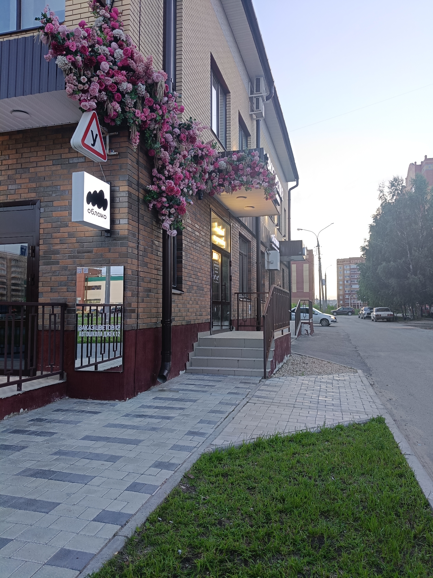 This media file depicts the exterior of a building adorned with abundant floral decorations, predominantly pink and white roses, cascading from the upper balcony and wrapping around a corner. The building itself is constructed of brick and features large windows and a prominent sign with the Cyrillic word "облако" (cloud) and a stylized cloud graphic. Below this sign, a rectangular mirror is mounted on the wall, reflecting the opposite buildings and the street. There is also a triangular road sign with the Cyrillic letter "У" inside, indicating a pedestrian crossing or a similar traffic rule.
The scene is set on a street in Yoshkar-Ola, Russia, as suggested by the location context. The pathway leading to the building is paved with interlocking concrete blocks, and a patch of green grass borders the walkway. Several cars are parked along the street, and a tall residential building can be seen in the background, along with trees.
The time of day appears to be late afternoon or early evening, with the sky showing a clear, pale blue hue, indicating fair weather. The lighting suggests the sun is low on the horizon. No people are visible in the image, creating a sense of quiet and stillness. The overall impression is one of a well-maintained commercial or residential area, with a touch of natural beauty provided by the extensive flower arrangements. Text visible includes the signage on the building, and potentially some text reflected in the mirror, though it is difficult to read clearly.