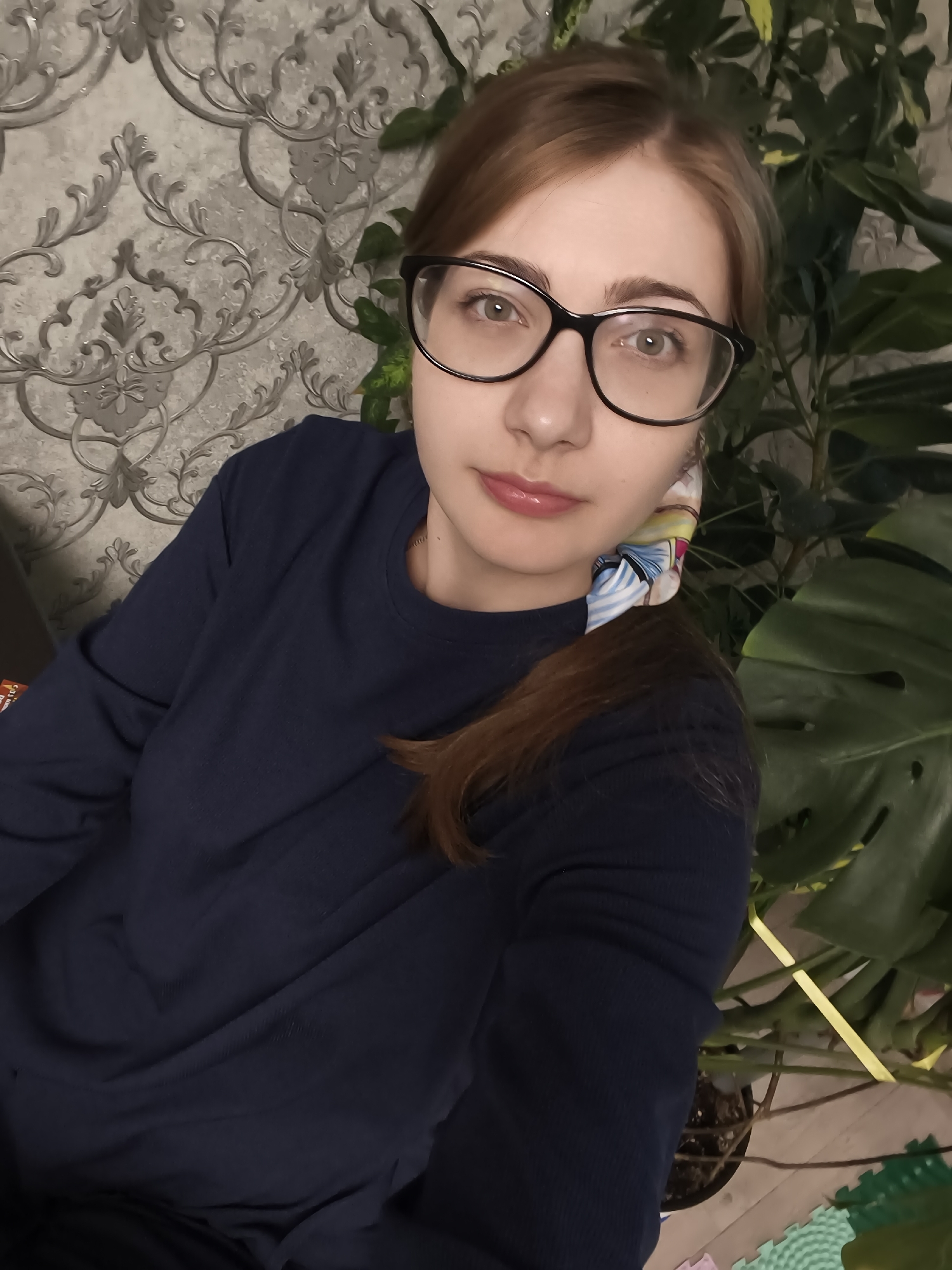 A young woman with light brown hair, wearing glasses and a dark blue long-sleeved shirt, is pictured in a selfie. She has fair skin and light green eyes. A colorful scrunchie with a pattern of cartoonish figures is visible in her hair, which is pulled back into a ponytail. She is wearing subtle makeup, with a touch of lipstick.
The background features a patterned wallpaper with a grey floral design. To her right, there is a large green plant with broad leaves, possibly a Monstera. The lighting suggests it is indoors, likely during the daytime. The overall mood appears casual and relaxed. The setting is consistent with a home environment.
There is no visible text in the image. The location context is Novaya Usman, Russia.