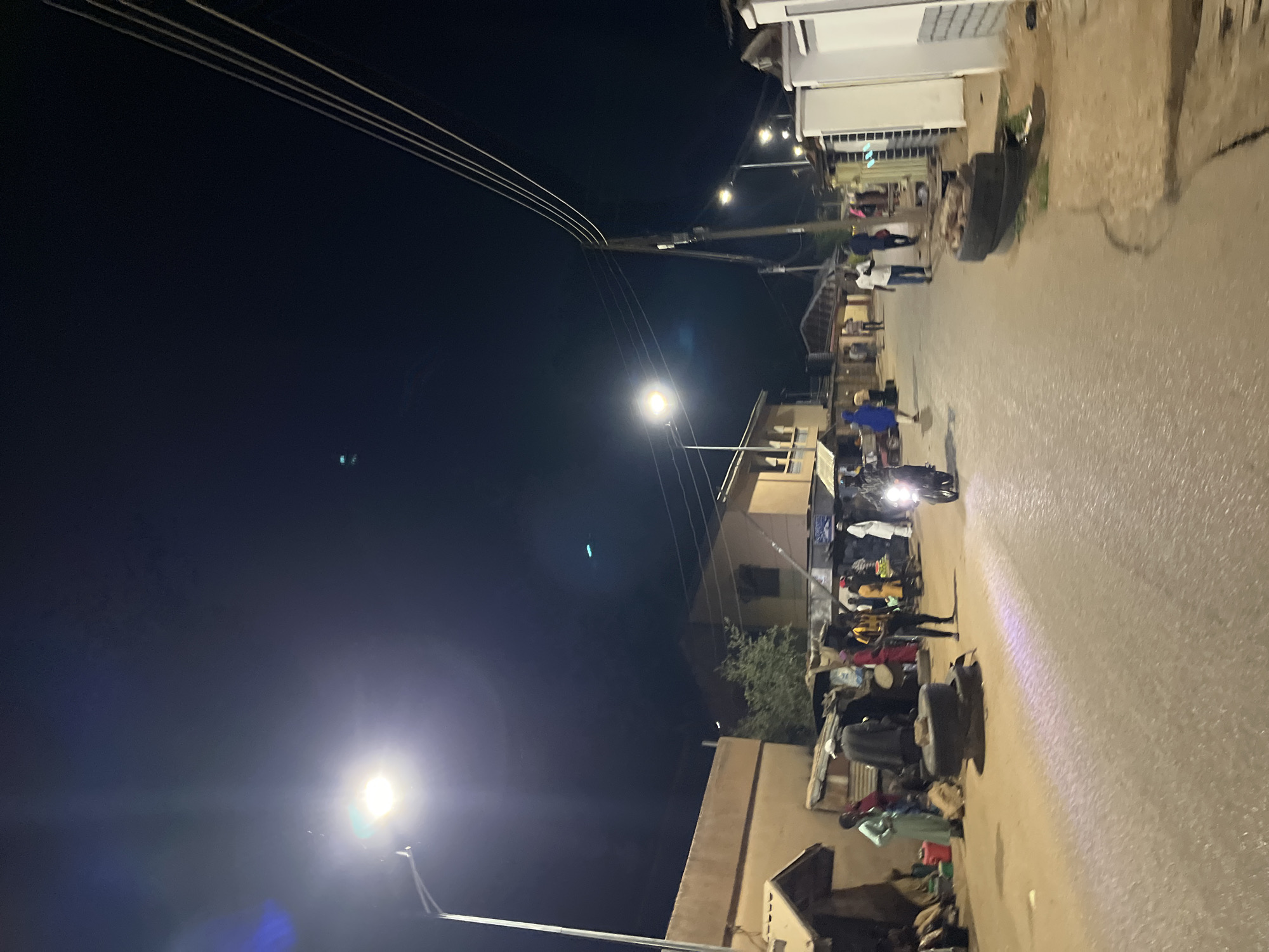 This night-time street scene, captured with a vertical orientation, depicts a busy urban or semi-urban environment in Jos, Nigeria. The dark sky indicates it is after dusk, with artificial lights providing the primary illumination.
A paved street, wet in places and reflecting light, runs through the center, lined on the right by various buildings that appear to be shops or residences. Overhead, multiple utility lines are visible against the dark sky. Several bright streetlights cast strong glows and lens flares, with one prominent in the foreground and others receding down the street.
Numerous people populate the street, primarily gathered on the right side. They appear to be engaged in various activities, suggesting a lively evening street scene, possibly a market or social gathering. A motorcycle with its bright headlight on is visible in the middle ground, surrounded by a cluster of individuals. Other people are seen standing, walking, and interacting along the thoroughfare. On the ground near the buildings, discarded tires and other objects, possibly goods or personal belongings, are scattered. No visible text is present in the image. The overall impression is one of an active community maintaining a vibrant presence on the street after dark.