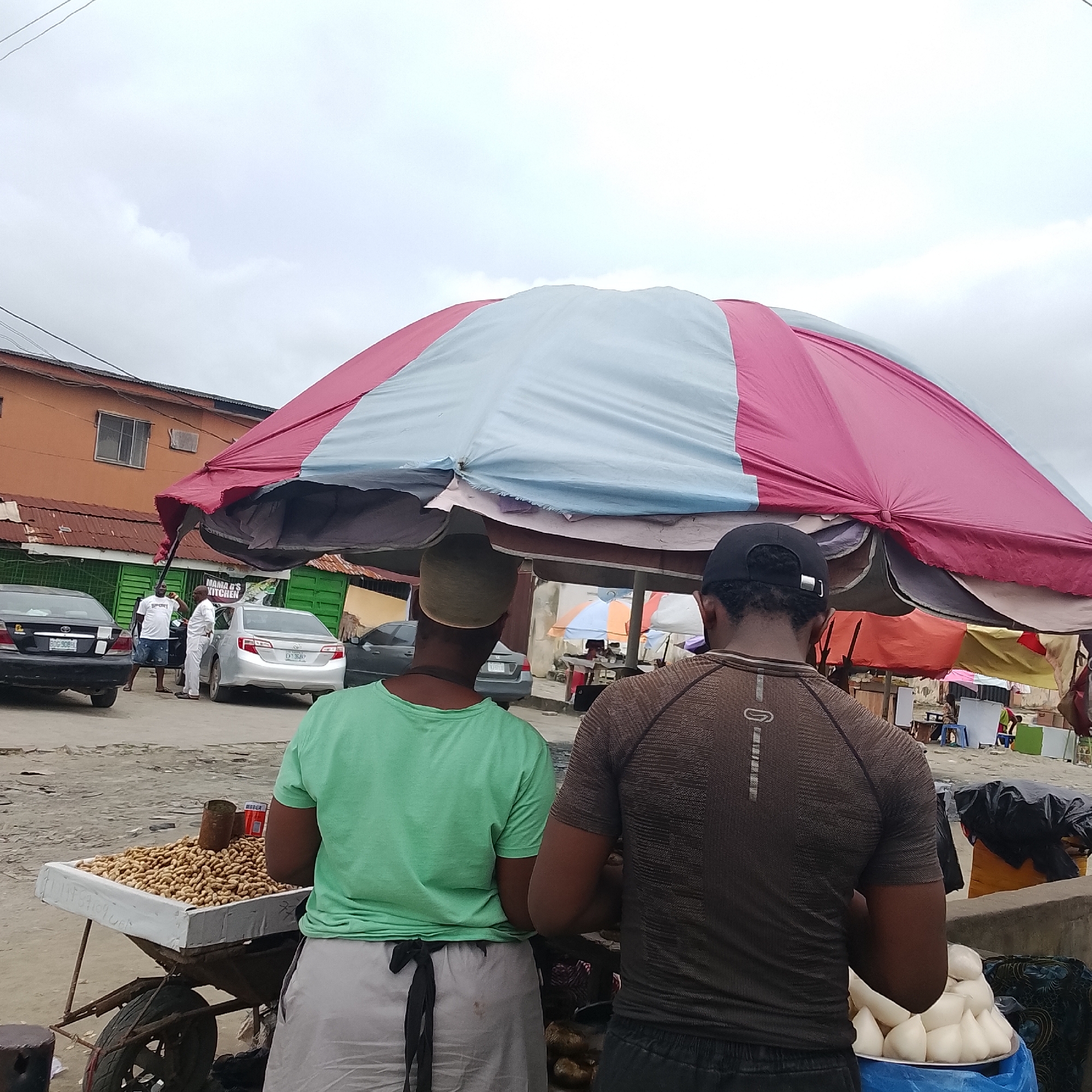 The image depicts an outdoor market scene in Ado, Nigeria, under an overcast sky. Two individuals are standing in the foreground, viewed from behind. The person on the left wears a light green t-shirt and a light-colored wrap tied at the waist. They are standing next to a cart filled with peanuts, with a metal can resting on top of the nuts. The person on the right wears a dark brown t-shirt and a black baseball cap. They are standing near a pile of white, rounded food items, possibly dumplings or yam preparations, on a blue platter. A large, multi-colored umbrella, with sections of pink and light blue fabric, covers a portion of the market.
In the background, several cars are parked, including a black sedan with a license plate ending in "BGH 90BL" and a silver sedan with a license plate ending in "DKJ 3549". Buildings with orange and green facades line the street, with corrugated metal roofs visible. A sign on one of the buildings reads "MAMA'S KITCHEN". Other market stalls with various umbrellas are visible further in the background. The ground appears to be a dirt or paved surface, characteristic of a bustling urban marketplace.