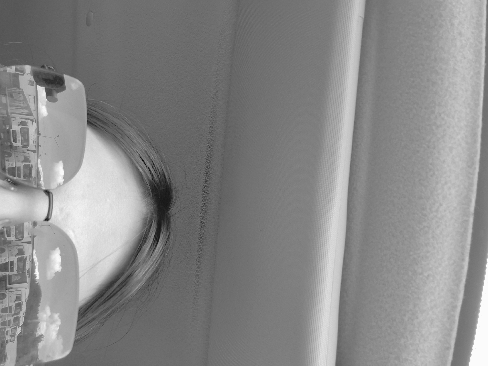 A grayscale, vertically oriented close-up photograph captures the upper portion of a person's head and the surrounding indoor environment. The subject's forehead and the top of their head are visible in the lower left section of the frame (if viewed with the head upright). Light-colored hair, appearing to be styled with a middle part or swept back, is clearly visible, along with a dark, thin hair tie or band around the head.
A prominent feature is a pair of rectangular eyeglasses worn by the person. The lenses brilliantly reflect an outdoor urban or suburban scene, showing multiple buildings resembling houses or apartment blocks, set against a bright, cloudy sky. This reflection indicates that the person is likely indoors, possibly looking out a window during the daytime in Izhevsk, Russia.
The background consists of various interior elements. To the right of the person's head, a textured wall with a subtle, rough pattern is visible. Further to the right, two different types of window coverings are present: a section of vertical blinds with distinct parallel lines, and an adjacent, softer, heavily textured fabric curtain or drape. The composition emphasizes the contrast between the detailed indoor setting and the reflected exterior view. No text is visible in the image.