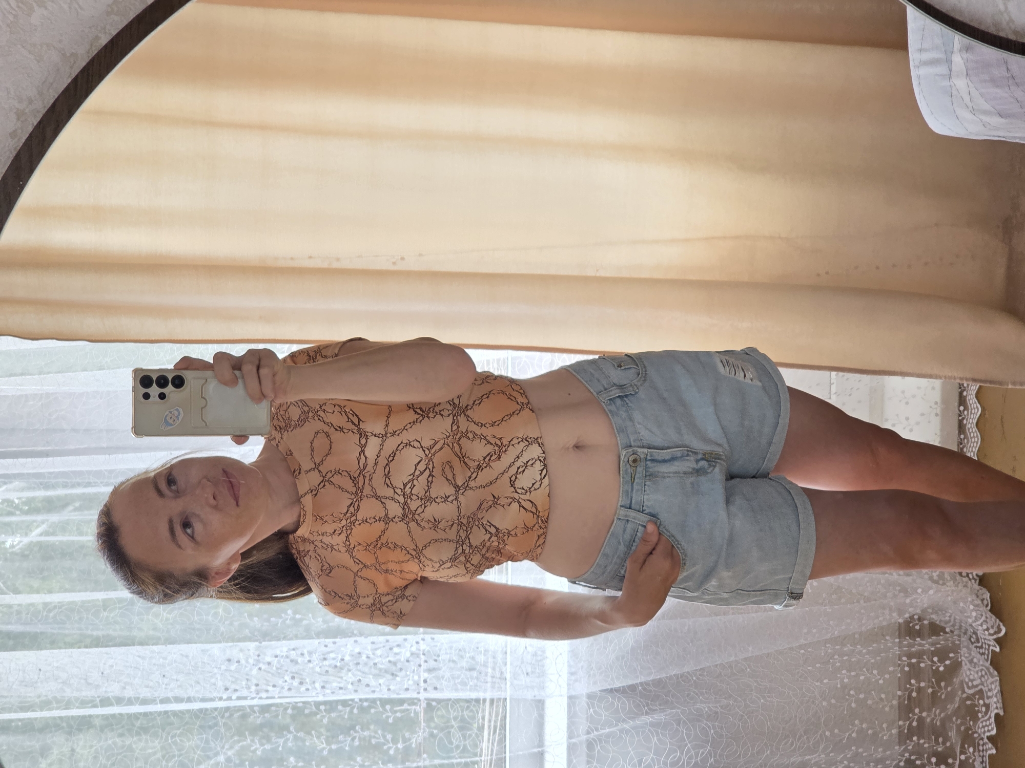 A woman with brown hair pulled back in a ponytail is taking a selfie in a mirror. She is wearing a cropped, short-sleeved, orange shirt with a black chain-link pattern and light blue denim shorts with ripped details at the hem. Her left hand is in her pocket. She is standing indoors, in front of a sheer white lace curtain on the left and a peach-colored curtain on the right. The mirror's frame is visible. The environment suggests a residential setting in Chaikovsky, Russia.