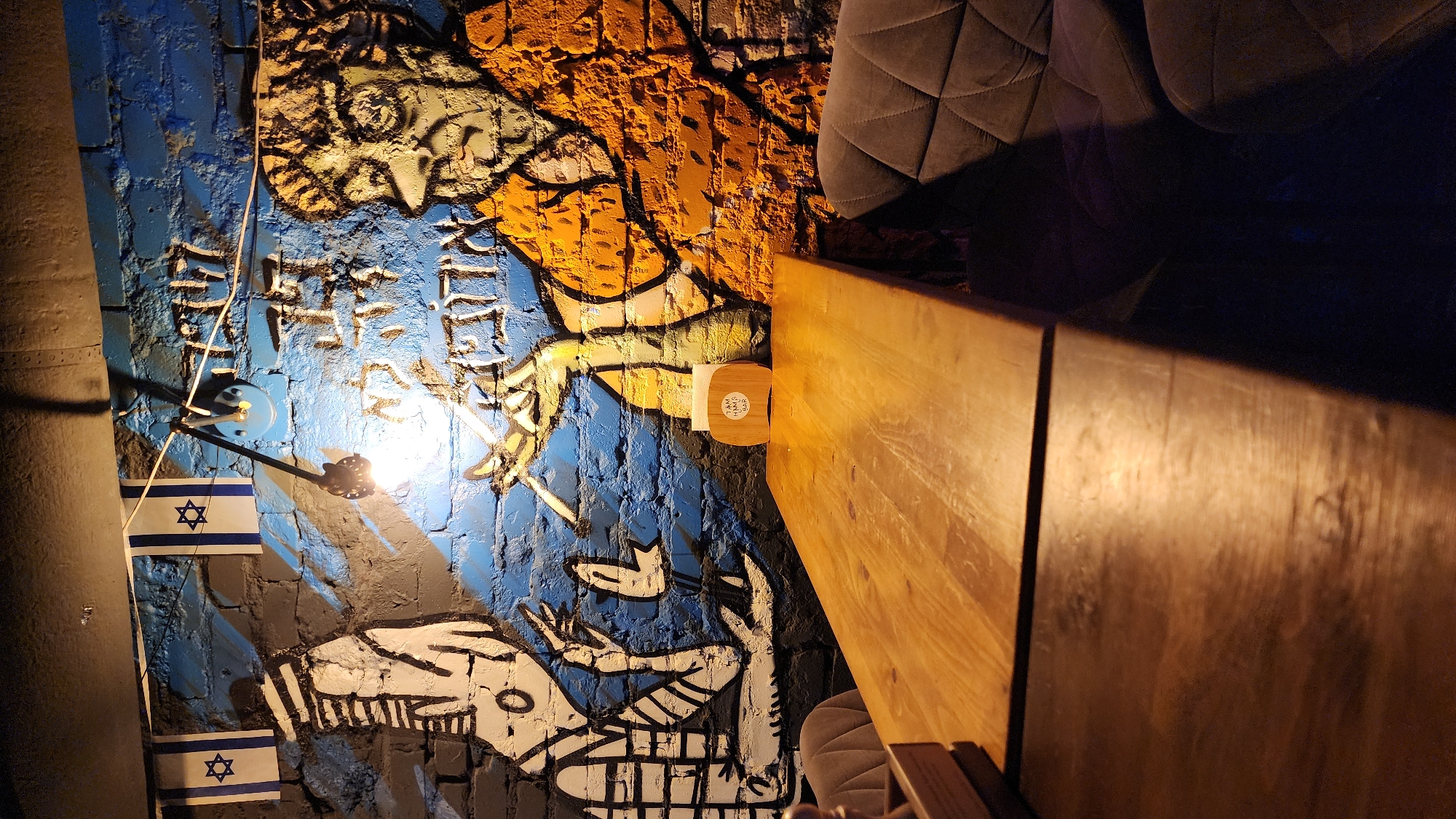 This is an interior photograph of a bar or restaurant located in Moscow, Russia. The scene features a distressed brick wall illuminated by a central, exposed incandescent light bulb mounted on a blue fixture. The wall is painted with vibrant murals: a dark grey section on the left displaying a stylized white figure with striped sleeves, a central blue section, and a right orange/red section featuring a cartoonish male figure with glasses and a beard, holding a cigarette or pipe. Above the figures, white Hebrew script is painted, reading "הלילה שלנו אדמתינו" (HaLayla Shelanu Admatenu), which translates to "The night is ours, our land." Two small Israeli flags, each depicting a blue Star of David on a white background with two horizontal blue stripes, are strung along a wire above the left side of the mural. A dark metal pipe extends horizontally across the top of the frame. In the foreground, a long, light-colored wooden table with visible grain occupies the center. A wooden napkin holder, engraved with "I AM HAM'S BAR" and a logo, is positioned on the table near the wall. Portions of two grey upholstered chairs with quilted backs are visible on the left and right sides of the table.