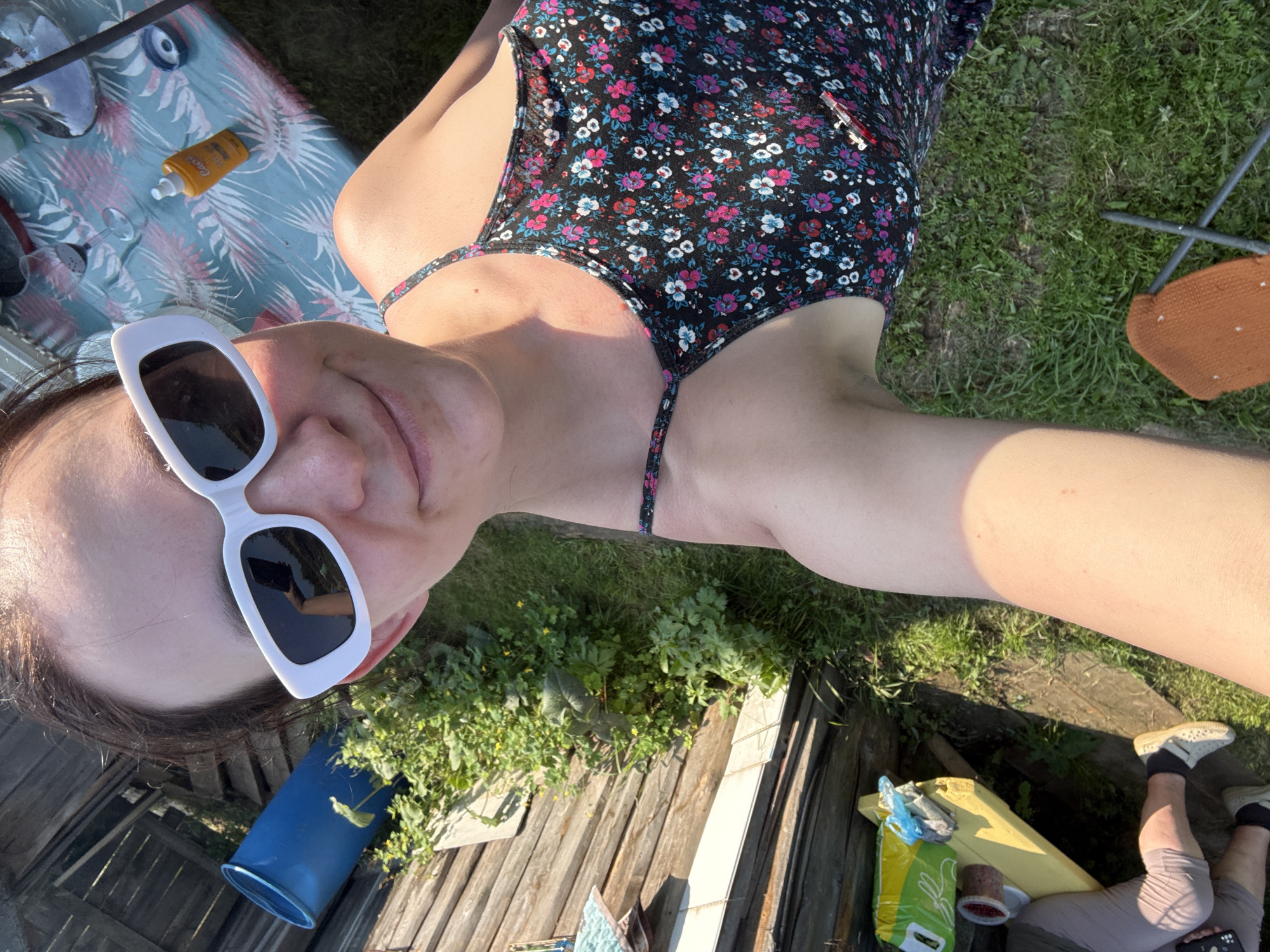 In a sun-drenched outdoor setting, a woman is captured in a selfie taken from a high-angle perspective, looking up towards the camera. She is wearing a floral-patterned dress and white, angular sunglasses. The scene appears to be a yard or garden, with lush green grass and wooden structures visible.
On a patterned mat or blanket to her left, there are a few objects including a small orange bottle, likely sunscreen, and what appears to be a drinking glass. Behind the woman, part of a blue container and some greenery can be seen. Further to the right, a small, orange, canvas chair with dark grey legs is placed on the grass.
The lighting suggests it is a bright, sunny day, with strong shadows indicating the time of day is likely mid-morning or afternoon. The overall mood conveyed is casual and relaxed, suggesting leisure time outdoors. The presence of sunscreen and the comfortable attire point towards an activity associated with warm weather, such as sunbathing or simply enjoying the outdoors. The background elements like the wooden fence and the general untamed greenery hint at a rural or suburban location.