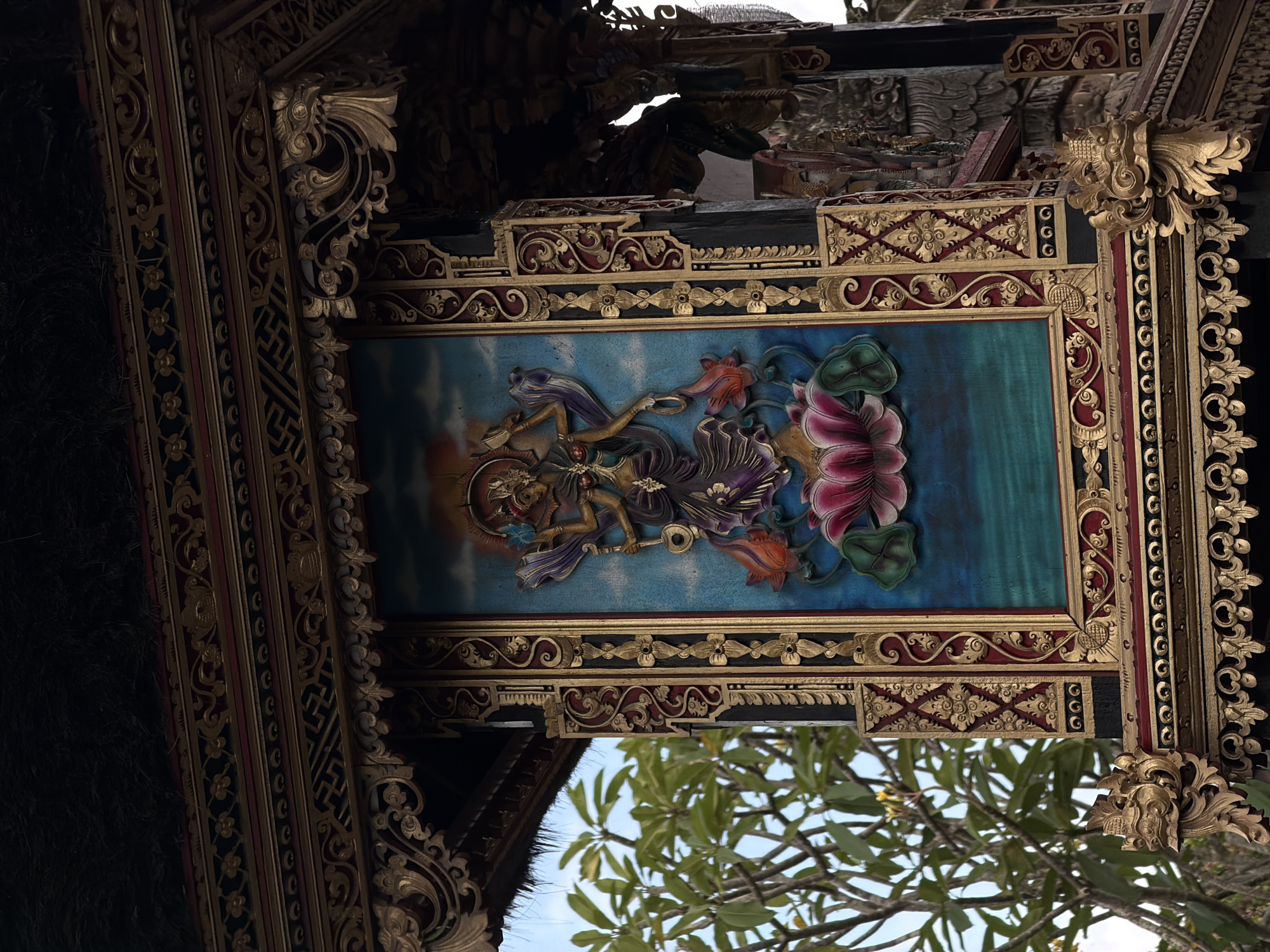 The image captures a section of a Balinese architectural structure, specifically a painted panel featuring Hindu deities and lotus flowers. The panel is framed with ornate carvings in gold and red, depicting intricate floral and geometric patterns. These carvings are part of a larger decorative framework, suggesting the ceiling or upper section of a building. Through the structure, greenery and a glimpse of the sky are visible. Based on the location information of North Kuta, Indonesia, it's likely the image depicts part of a temple or a traditional Balinese building, rich in cultural and religious symbolism.