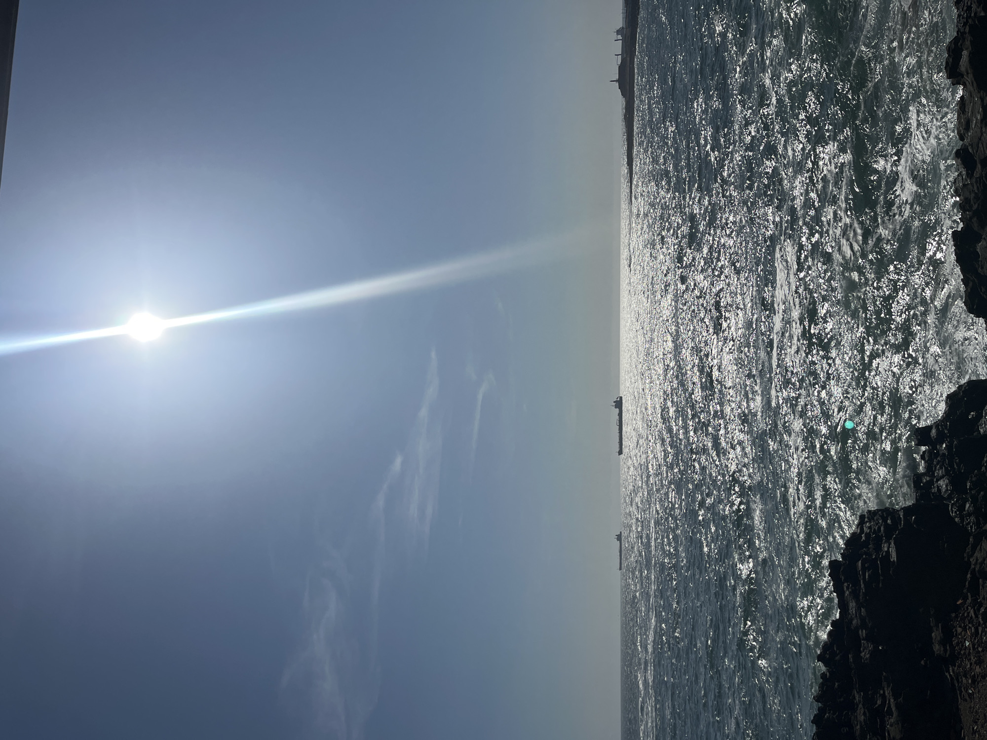 The image depicts a view of the Pacific Ocean from the coast of Arica, Chile. The sun is prominently positioned in the sky, emitting a strong glare. The ocean water is choppy and reflects sunlight. In the distance, there is a faint outline of the city's coastline and some structures. Two cargo ships are visible on the water. The foreground contains dark, rocky formations that form the coastline.