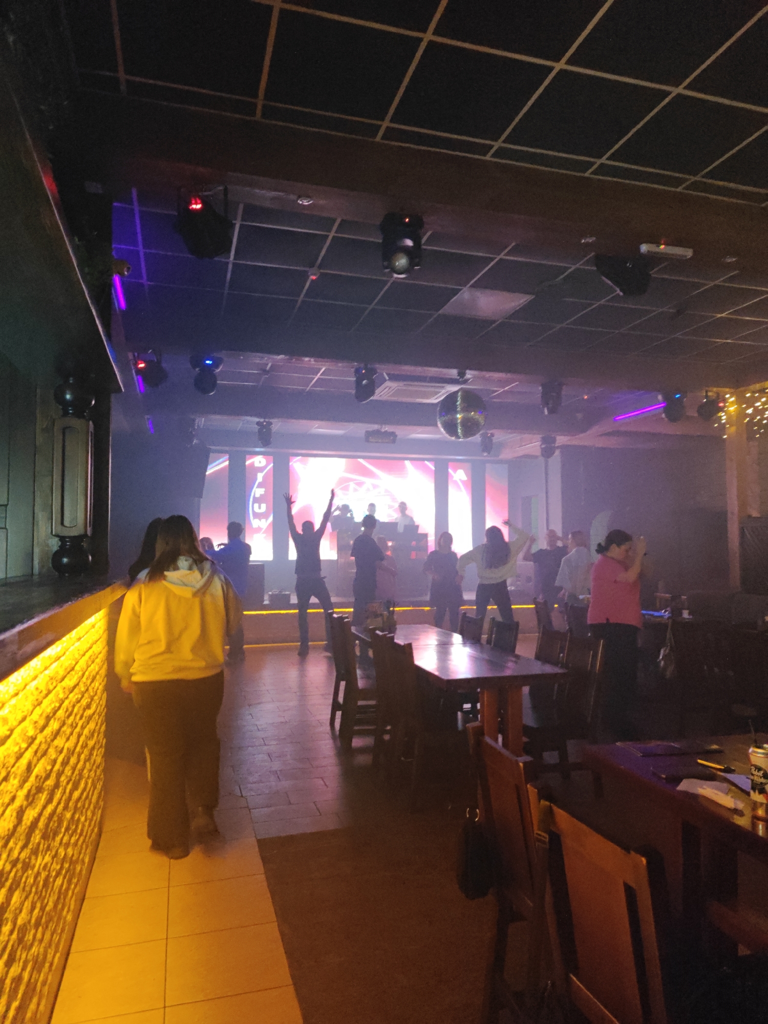 A brightly lit entertainment venue in Chaikovsky, Russia, features a crowd of people dancing near a stage with a lit display. A disco ball hangs from the ceiling amid numerous spotlights. Tables and chairs are situated throughout the space, some occupied, with drinks visible on one table. Two individuals are walking towards the dance floor in the lower left corner. The establishment appears to be a nightclub or bar with a dance floor.