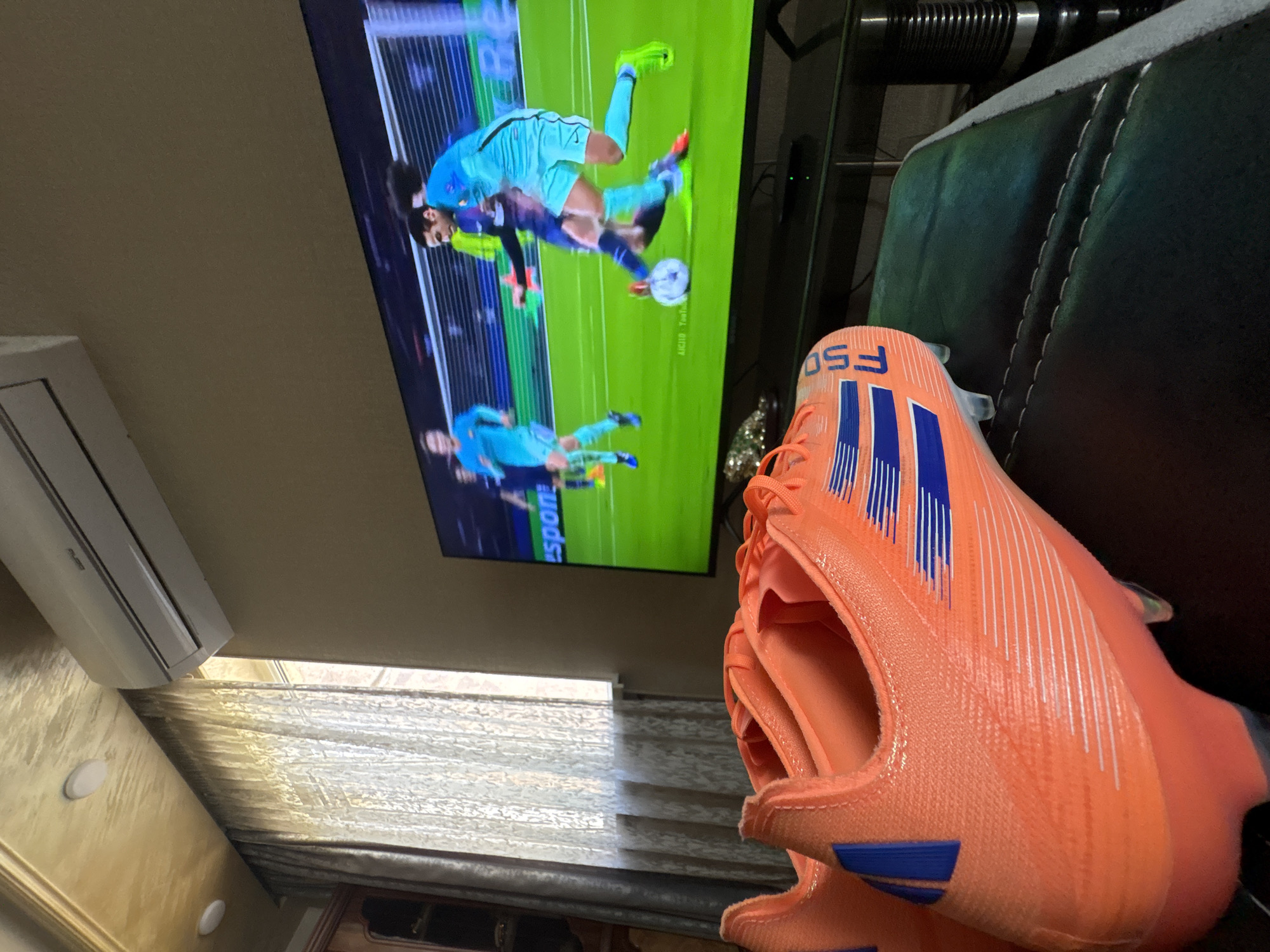 The scene depicts an indoor setting, possibly a living room in Yerevan, Armenia. An air conditioning unit is mounted on the wall near a window covered with horizontal blinds. A flat-screen television displays a soccer game between FC Barcelona and Paris St Germain. In the foreground, there is a pair of orange Adidas F50 soccer cleats. The background shows part of a black leather sofa.