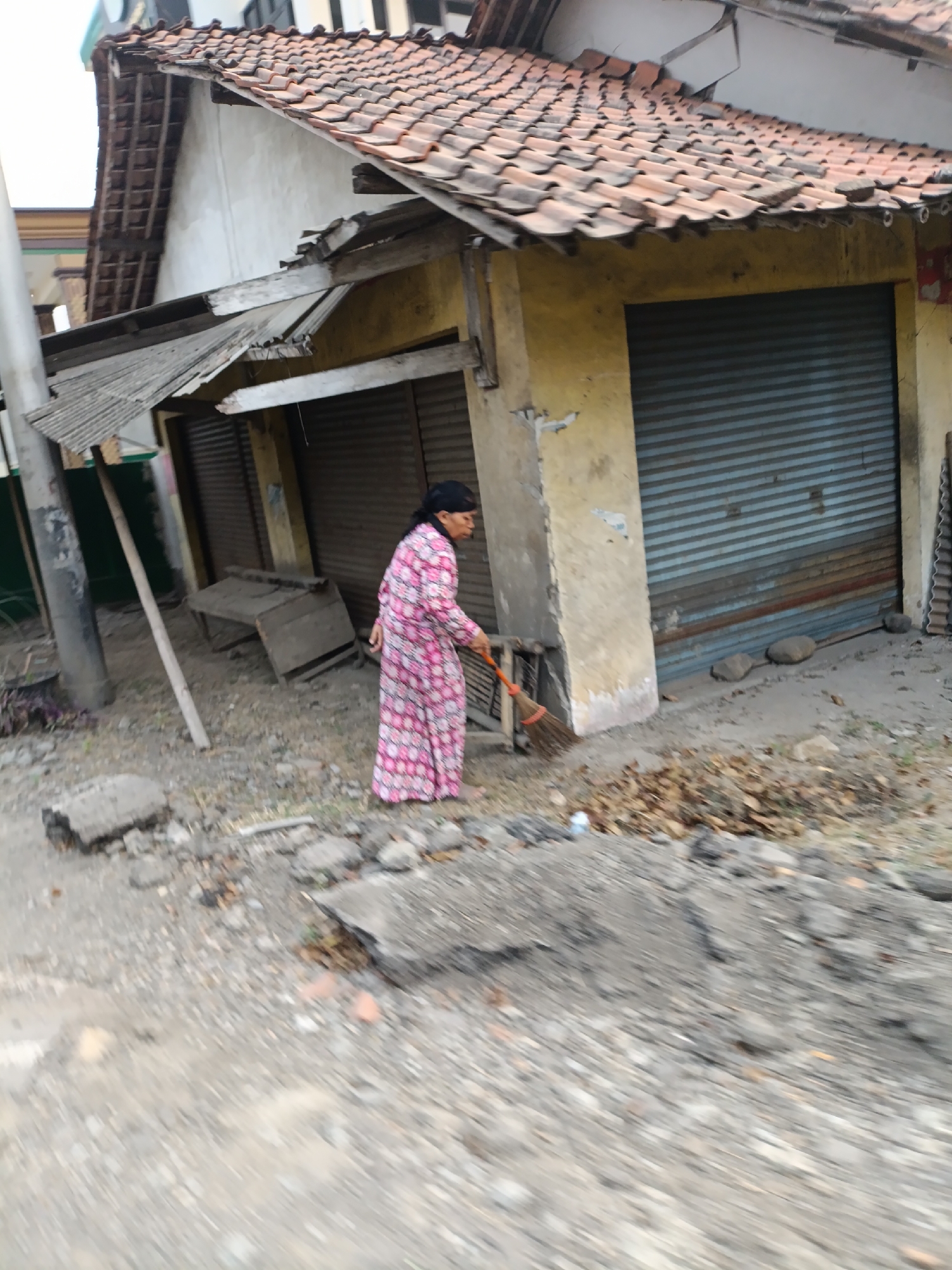 The image depicts a street scene in Cariu, Indonesia, focused on a rundown building. A woman, wearing a pink and white patterned dress and a headscarf, is actively sweeping a pile of leaves with a broom. The building itself shows signs of disrepair, with a weathered yellow facade, a partially collapsed corrugated metal awning, and a roll-up metal door. Debris, including rocks and broken concrete, litters the ground in front of the building. A rough, unpaved road or path is visible in the foreground. The overall environment appears to be an urban or semi-urban area, possibly a commercial district with shuttered storefronts.