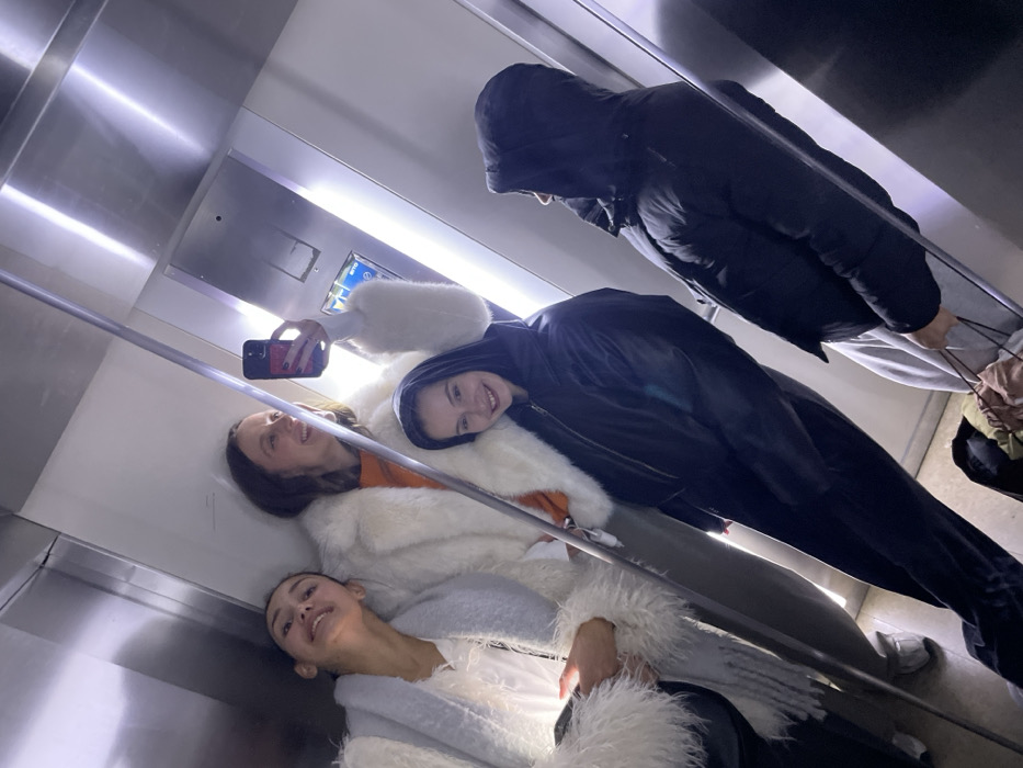 Inside a modern elevator, three young women pose for a selfie. One holds a phone, smiling with an orange top and white fuzzy jacket. Another, also smiling, wears a black hooded jacket over a white jacket. A third woman in a similar white fuzzy coat looks up. An additional person in a black puffy jacket and gray pants stands near the back, facing away from the camera. The elevator's interior is brightly lit with reflective silver walls.