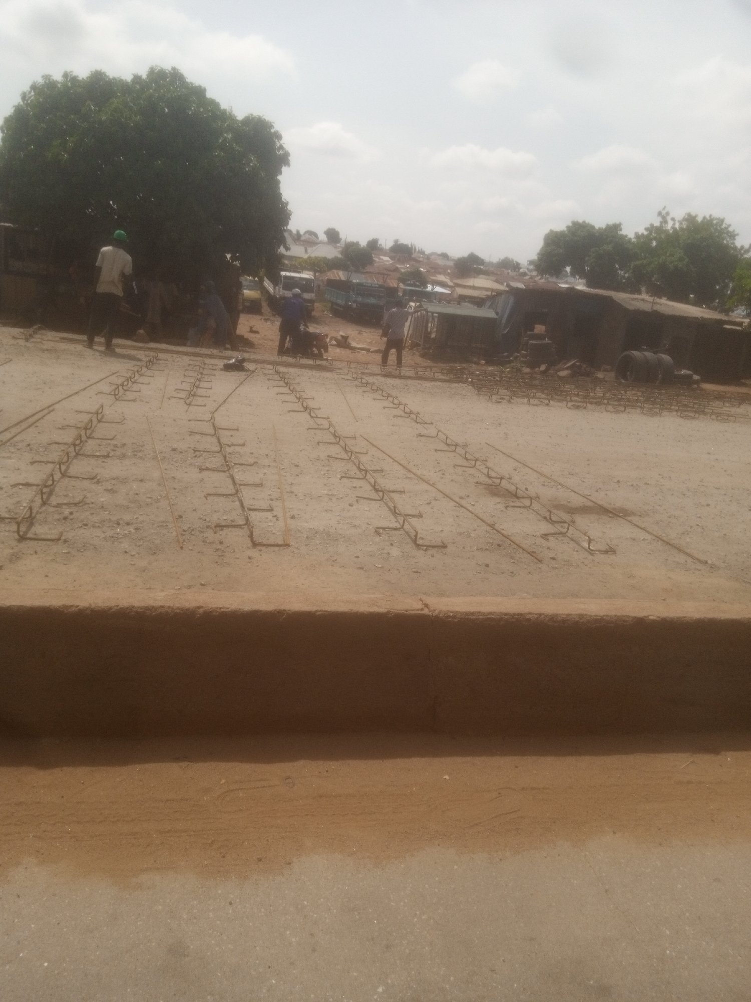 The image captures an outdoor construction site in Nassarawa, Nigeria, under a cloudy sky. The foreground features a road with a raised dirt embankment. Beyond this, a vast expanse of sand is covered with rows of rebar, arranged in a grid pattern, indicating preparation for a concrete pour. In the midground, several men, some wearing hard hats and work clothes, are visible, engaged in construction activities. There are also various vehicles, including trucks and what appear to be construction equipment, as well as some rudimentary structures and trees. The background shows a dense settlement of buildings, typical of an urban or semi-urban African environment. The overall scene conveys ongoing infrastructure development.