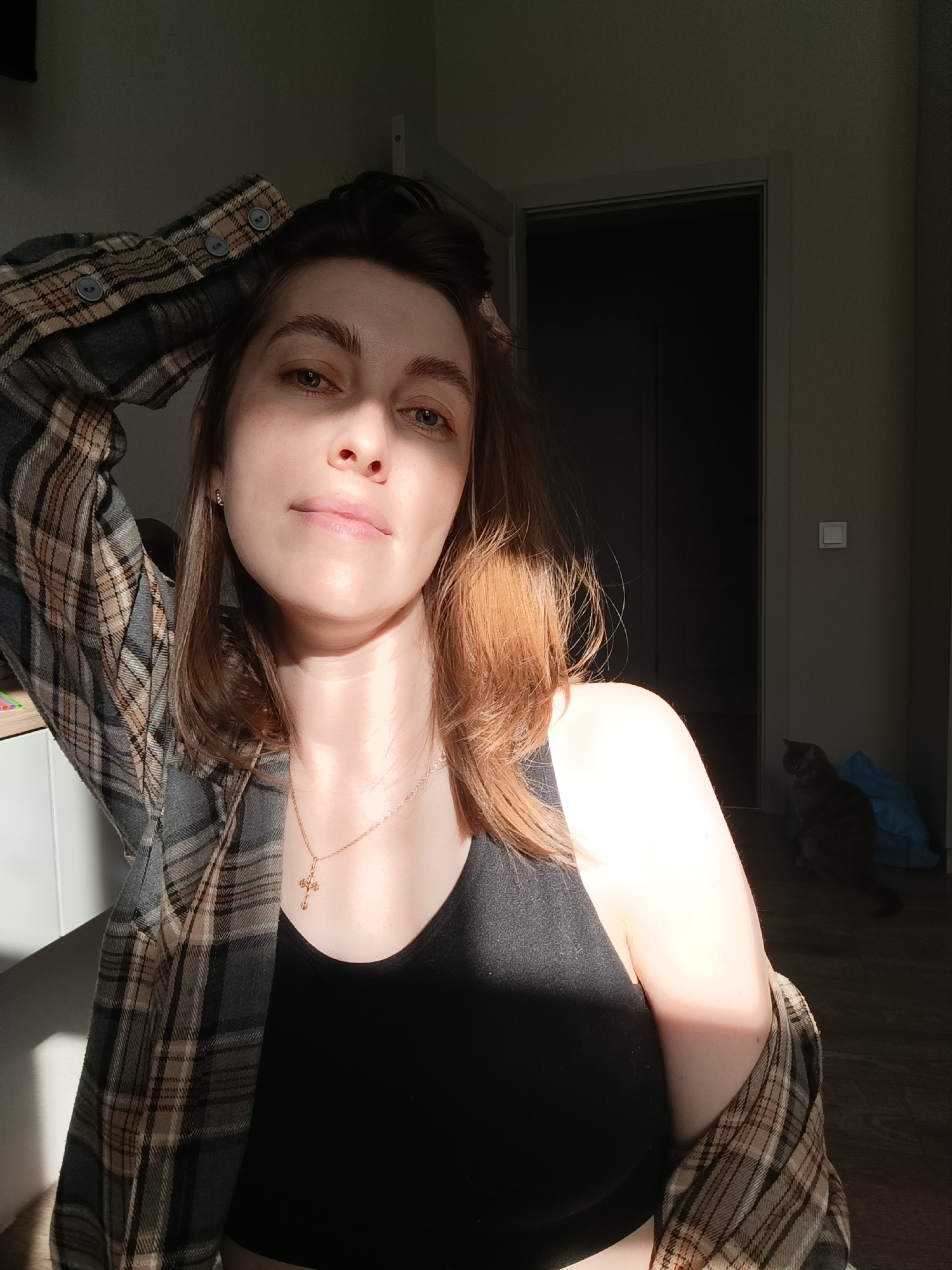 A woman is in a room lit by sunlight. She has brown hair and is wearing a black tank top and a plaid flannel shirt. She is wearing a gold necklace with a cross. Her left hand is in her hair. In the background, there is a doorway leading to a darker room, and a grey cat sitting on the floor. The photo appears to be taken indoors. There is no information about the location being in Chaikovsky, Russia.