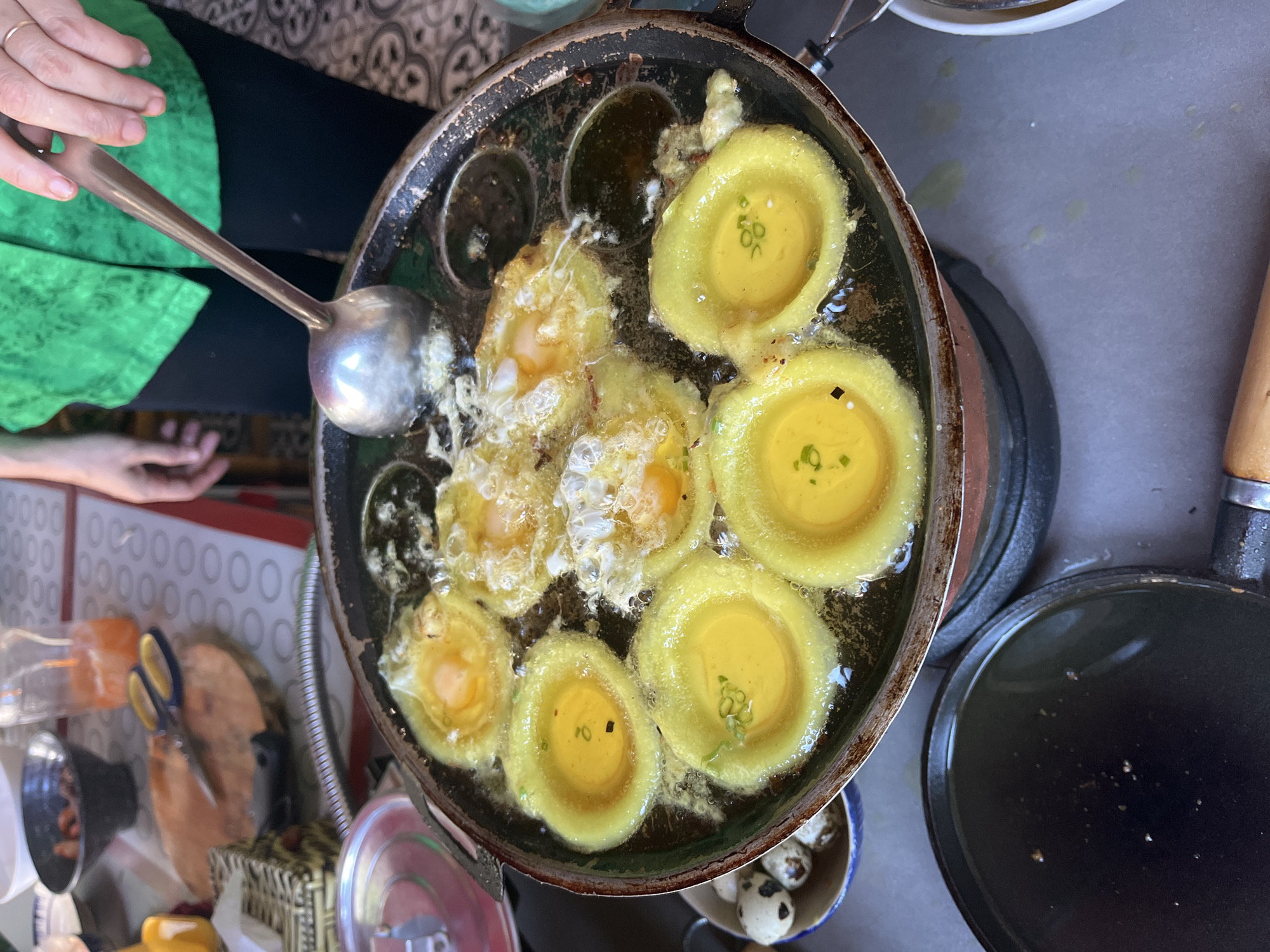 Overhead shot of a street food vendor in Đà Nẵng, Vietnam, cooking Bánh Khọt in a multi-sectioned cast iron pan on a portable stove. The pan contains multiple small cakes frying in oil, some with a solidified yellow outer ring and a runny yolk center, garnished with chopped scallions. A person with a green shirt is using a ladle to add oil. To the left are miscellaneous kitchen tools, a bowl of what appear to be quail eggs.