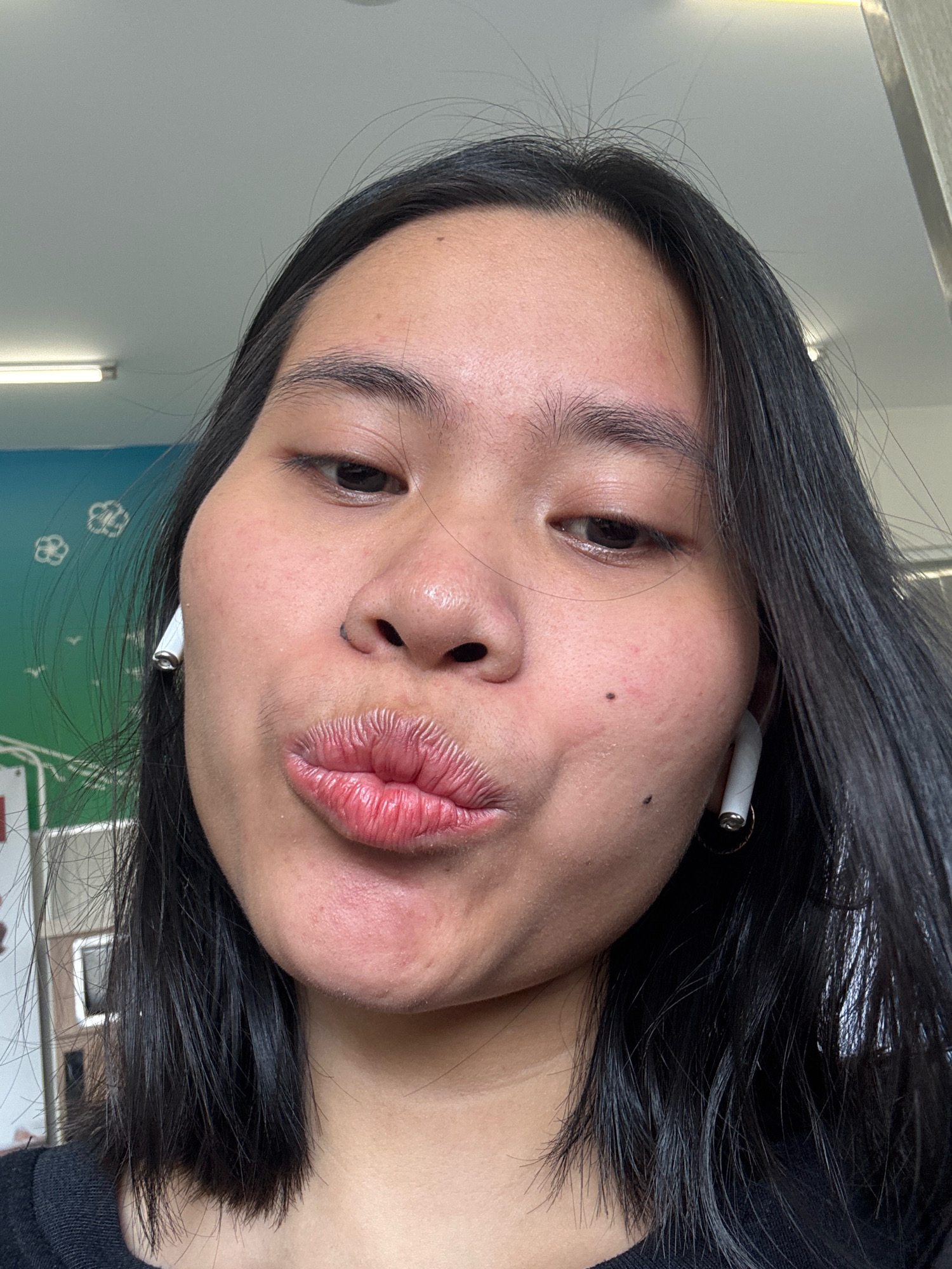A young Asian woman with black hair is taking a selfie. She is wearing white wireless earbuds and a black shirt. Her lips are pursed in a kiss pose. The background appears to be an indoor environment with a wall decorated with a mural of trees and clouds. The lighting suggests it is daytime. The location is East Jakarta, Indonesia.