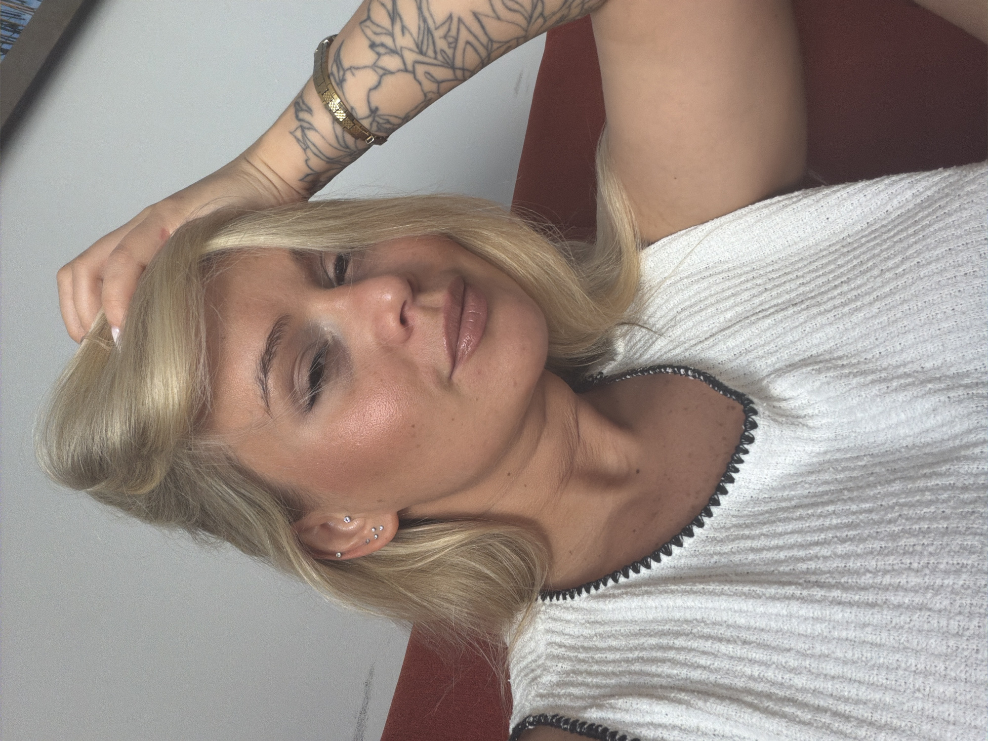 A woman with blonde hair, wearing a white top, is shown from the chest up. She has her left hand in her hair and her right arm is bent, resting near her head. She is wearing multiple earrings in her left ear and a gold bracelet on her right wrist. She has a tattoo on her right arm. The background is a light-colored wall and a hint of a red surface. The image is a close-up selfie. There is no indication of the city of Sarapul, Russia in the image.