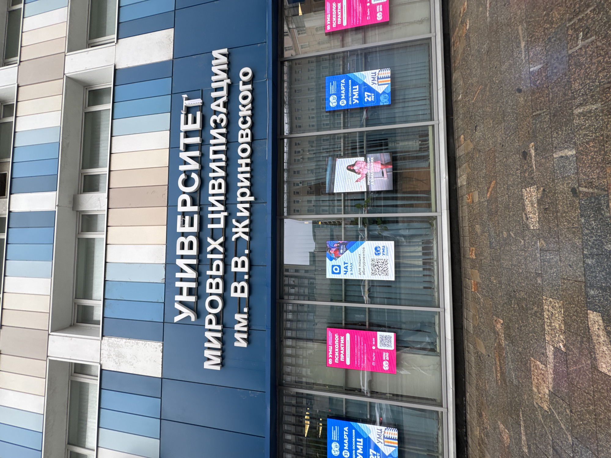 The image shows the exterior of the Moscow University of Civilizations, located in Moscow, Russia. The building features a blue facade with large windows. Several posters are displayed on the windows, advertising events such as a "Psychologist-Practitioner" course and a "CHAT for MAX" event for prospective students. The posters include dates, times, and QR codes. The street in front of the building is paved with granite tiles. The sky is overcast, suggesting a cloudy day.